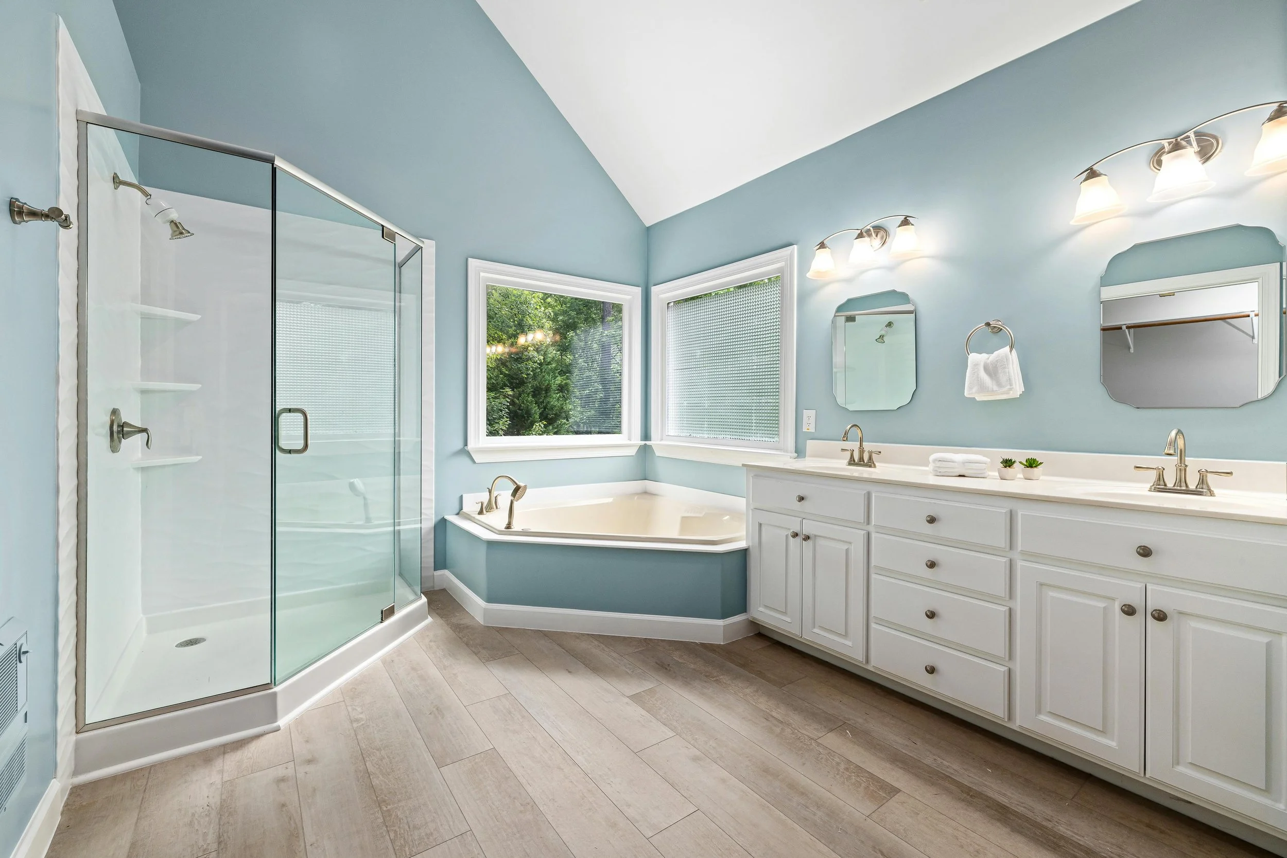 Bathroom with light blue walls, a window with lush green trees outside, a glass shower enclosure, a bathtub, a white vanity with a mirror, and lighting fixtures.