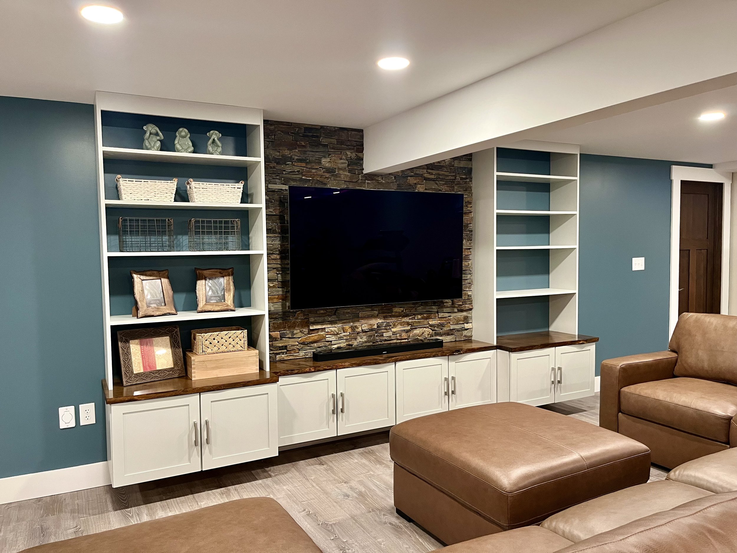 Living room with teal accent wall, white built-in shelves with decorative items, stone accent wall with a mounted television, brown leather ottomans, and a tan couch.