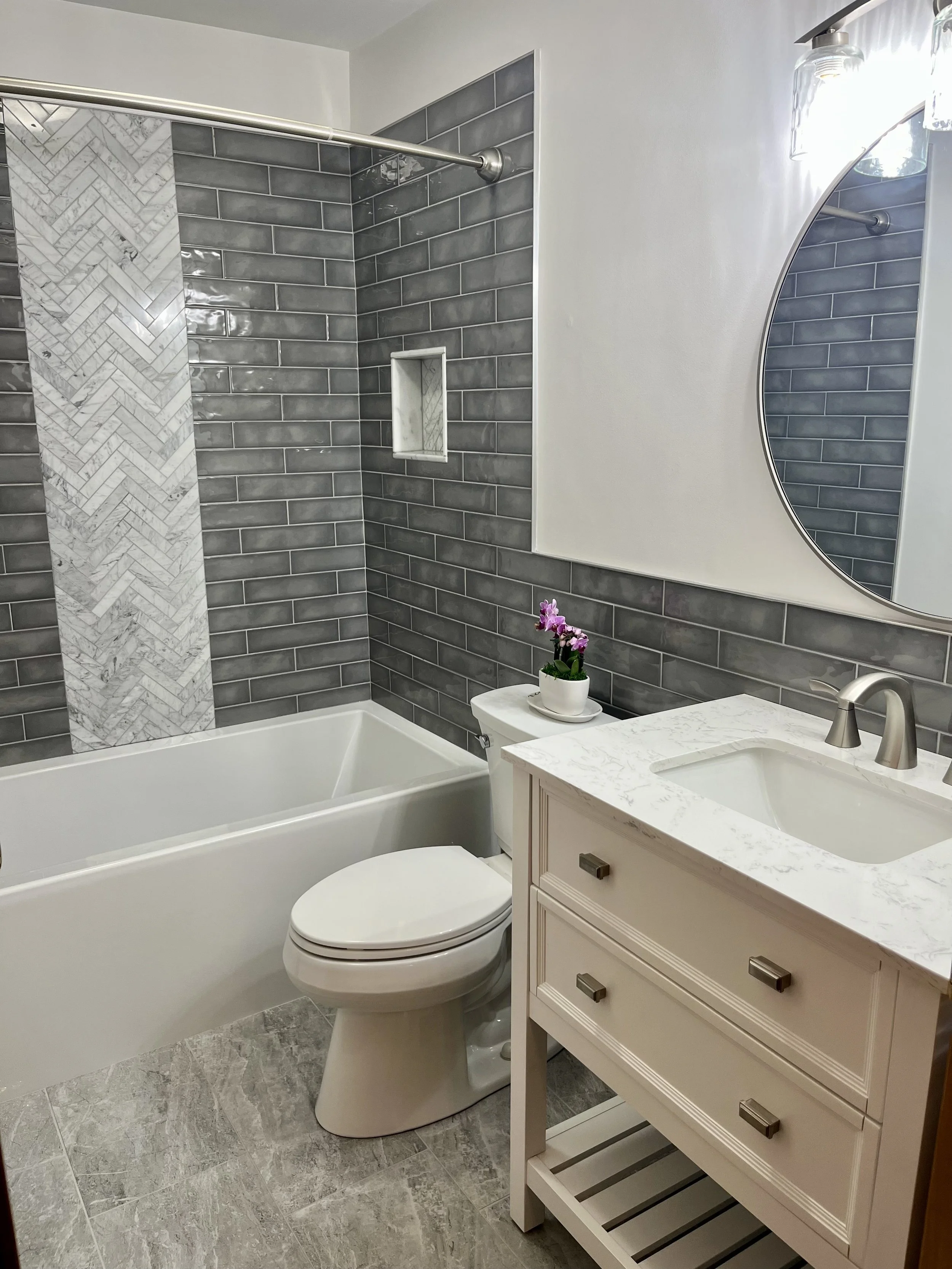 Bathroom with gray tiled shower, white bathtub, toilet, and a white vanity with a marble top and a round mirror.