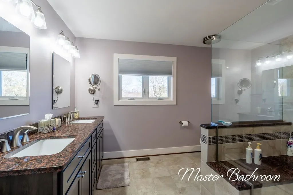 A spacious master bathroom with dual sinks, granite countertops, large mirrors, a window, and a glass-enclosed shower with tiled walls. Various toiletries are on the counters and inside the shower.