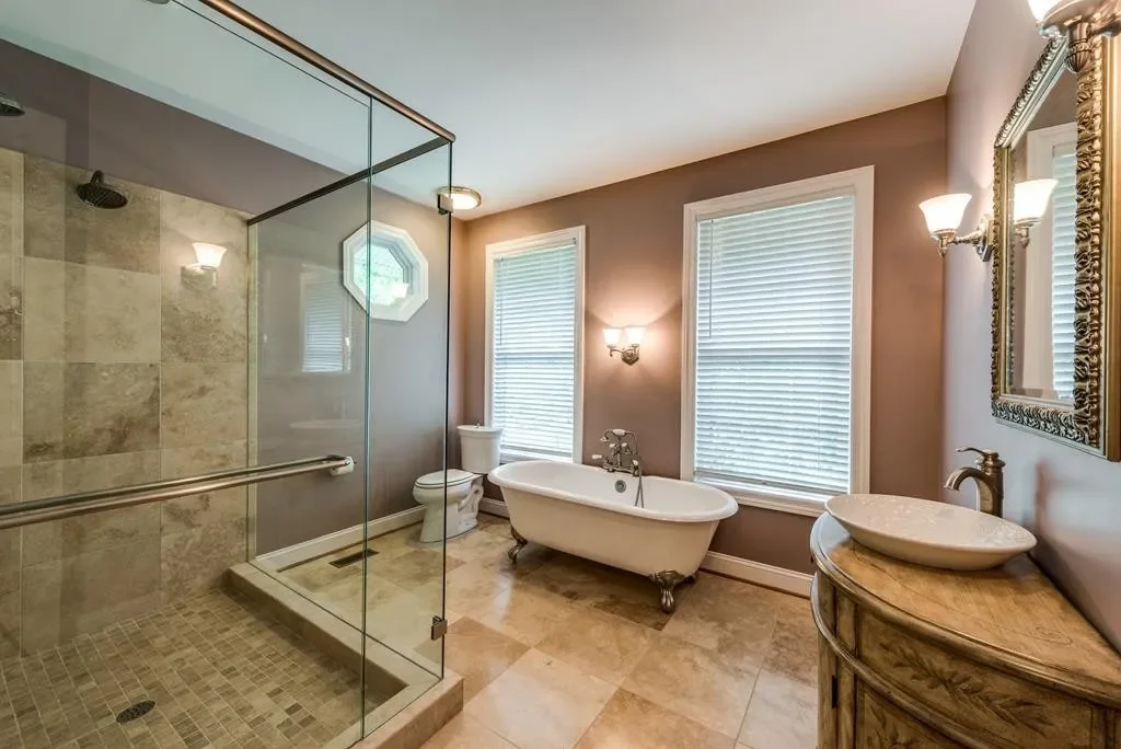 Bathroom with a glass-enclosed shower, clawfoot bathtub, toilet, pedestal sink, and decorative mirror, with beige tile flooring and beige walls.