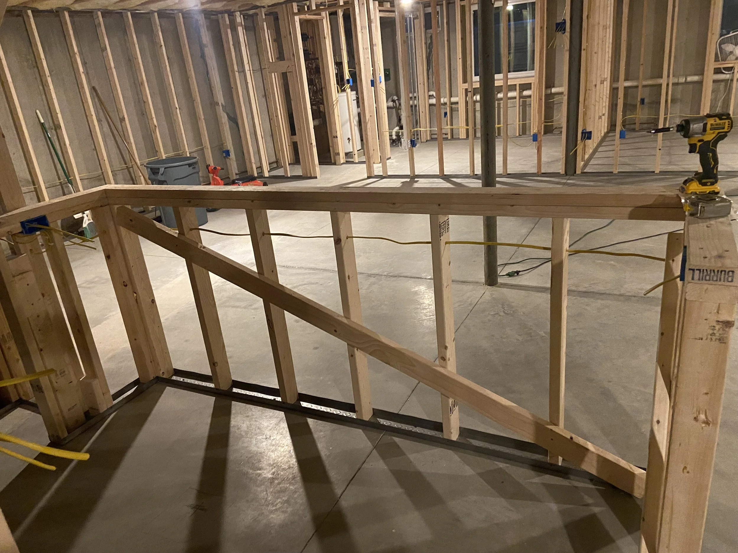 Interior of a house under construction showing exposed wooden framing, with construction tools and materials like a drill, wires, and a trash bin.