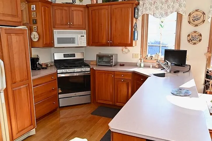Kitchen with wooden cabinets, white countertop, white microwave, stainless steel stove, small oven, and a window with curtains. There is a small TV and some kitchen appliances on the counters.
