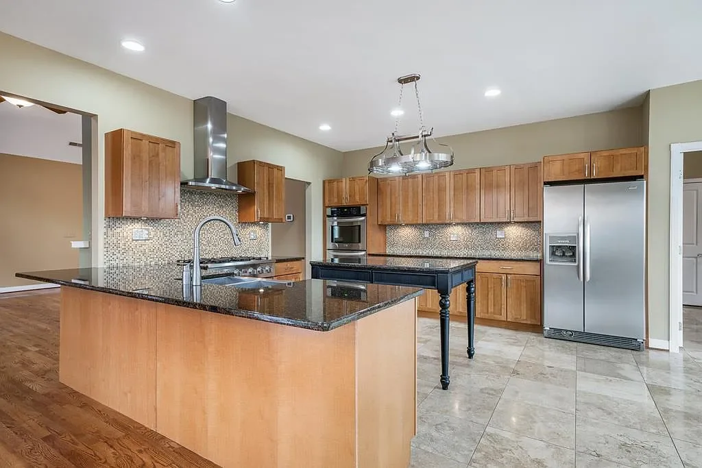 Modern kitchen with wooden cabinets, stainless steel refrigerator, black granite island, mosaic tile backsplash, and a central light fixture.