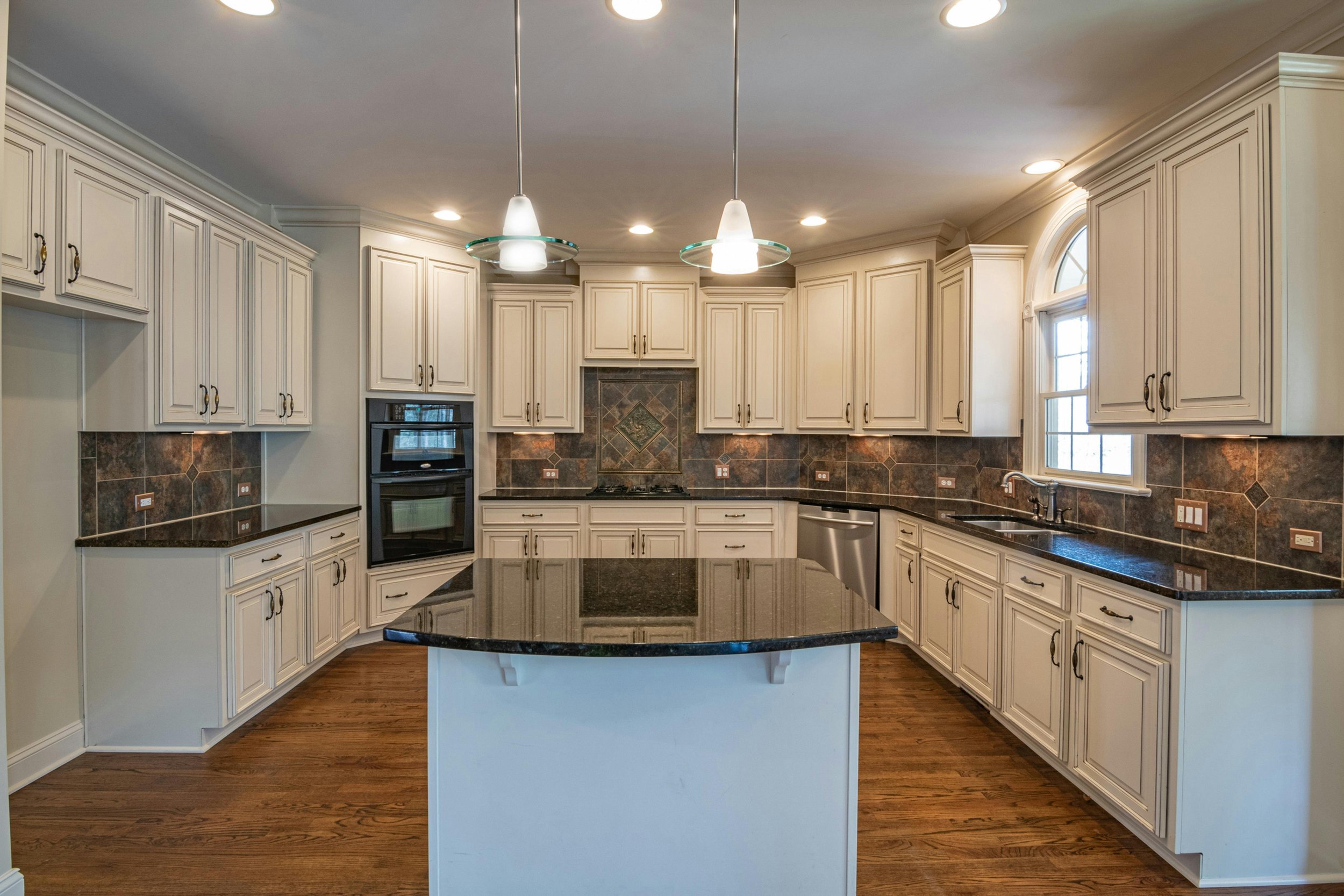 Kitchen with white cabinets, dark granite countertop island, built-in oven, stove, and double sink near window, with hardwood floors and pendant lights.