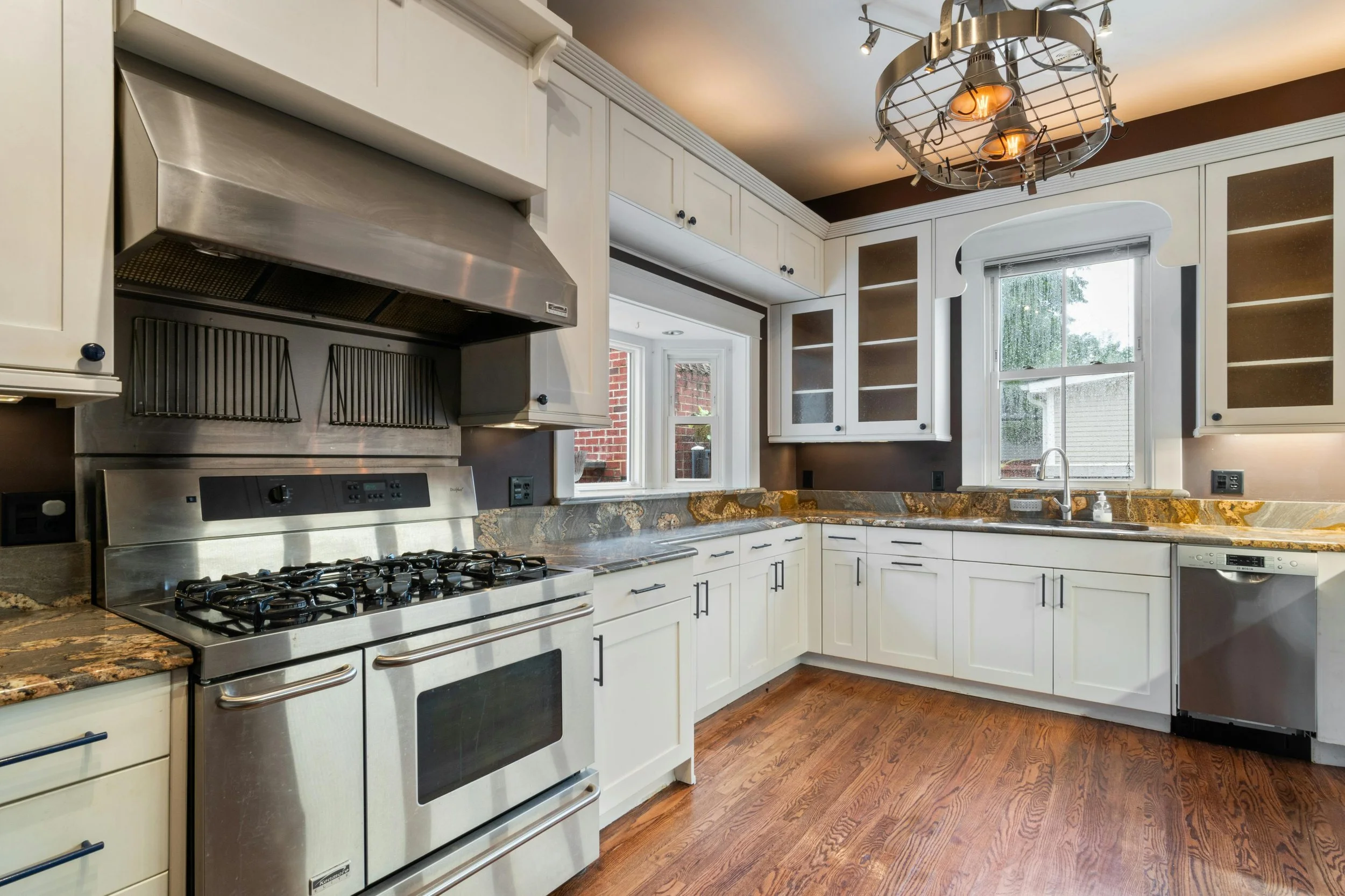A kitchen with white cabinets, stainless steel stove, granite countertops, and large windows, illuminated by an industrial-style ceiling light.