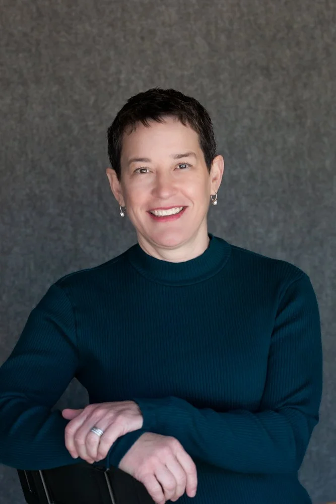 A woman with short dark hair smiling, wearing a dark sweater, earrings, and a ring, standing against a gray textured background.