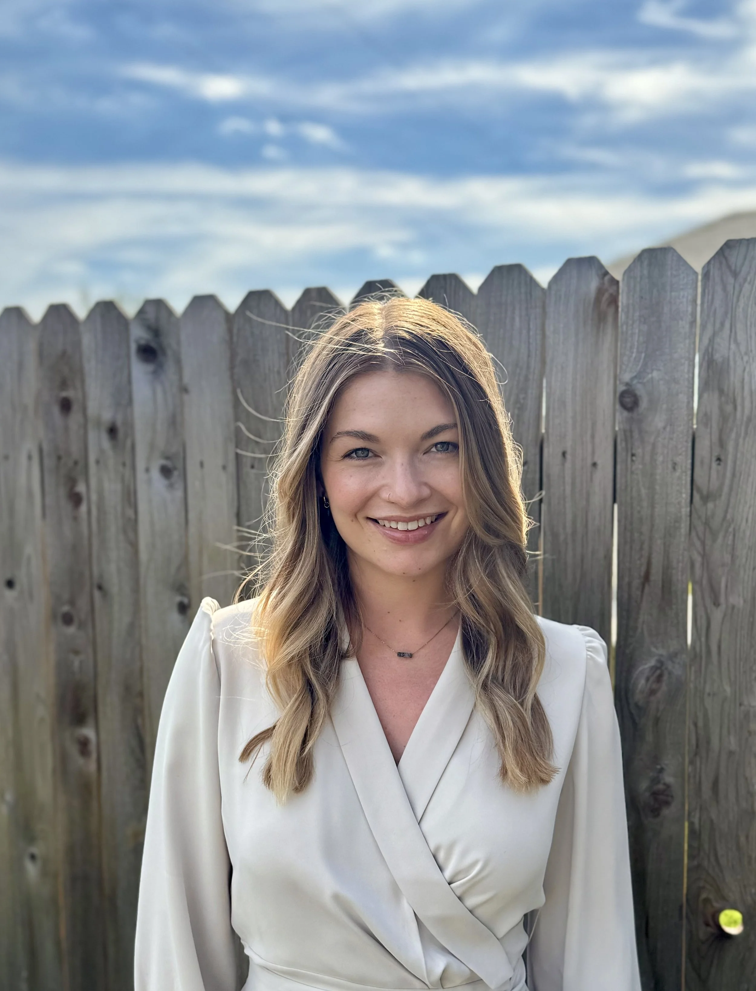 A woman with long, wavy blonde hair and blue eyes smiling, wearing a white wrap blouse, standing in front of a wooden fence under a cloudy sky.