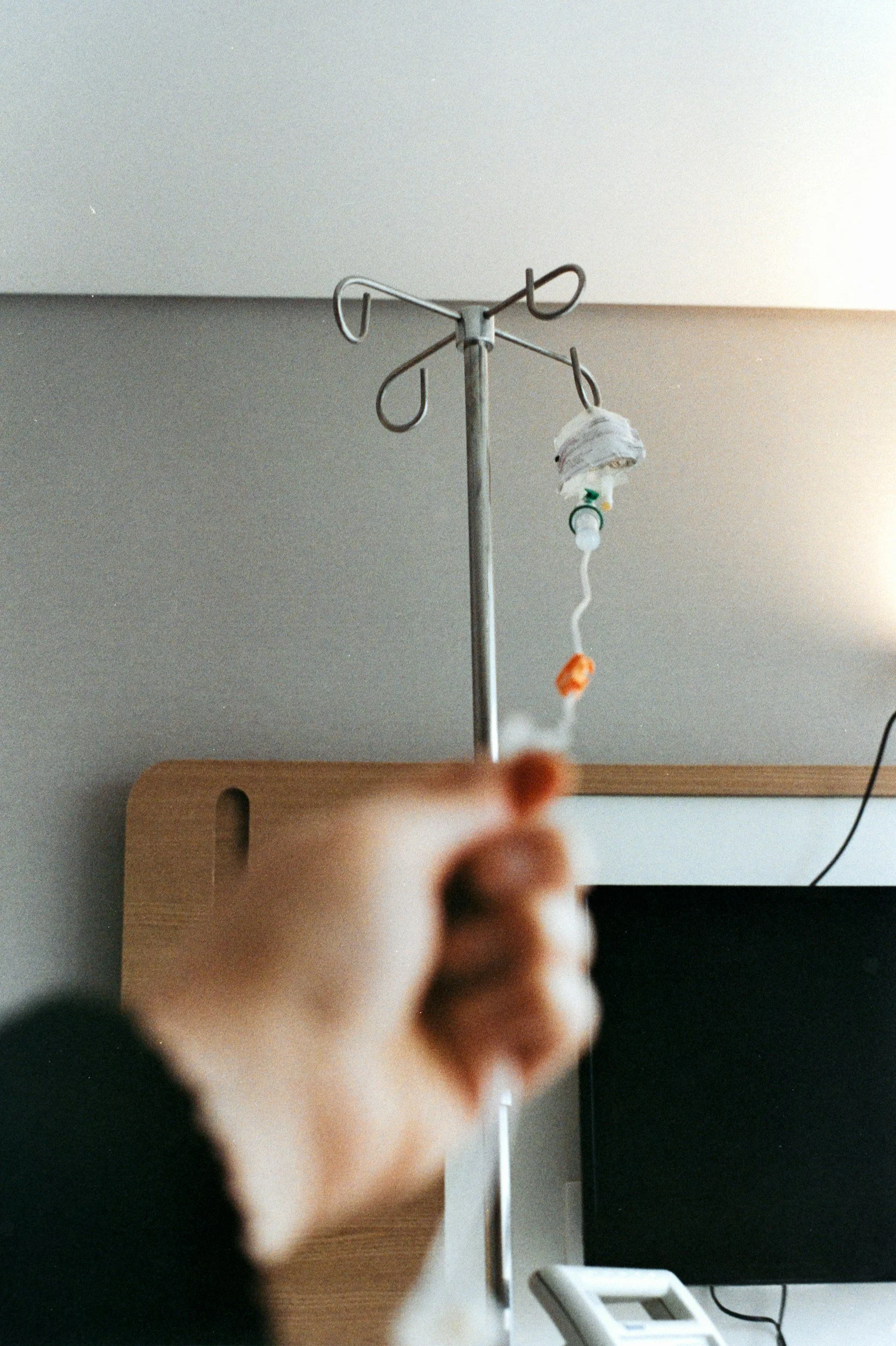 A hand reaching towards an IV pole with medical equipment attached, in a room with a wall and a wooden surface.
