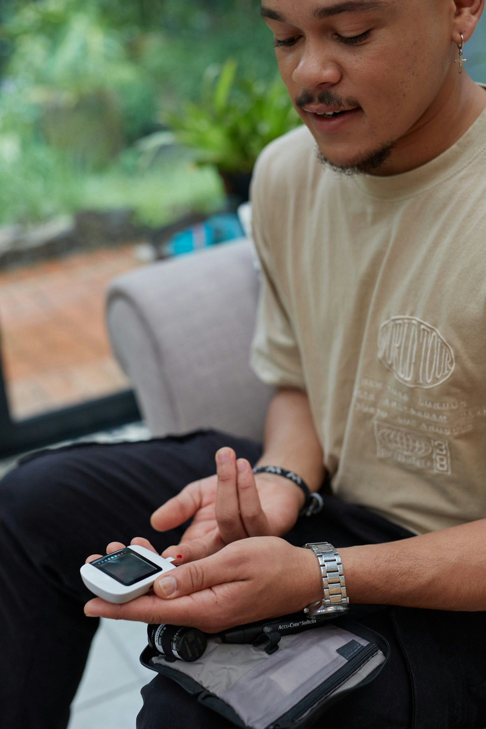 A man is sitting and looking at a glucose monitor displaying blood sugar level readings. He is wearing a beige t-shirt, a wristwatch, and has a thin beard and mustache. Another person's hand is giving him a fingerstick blood sample. The background appears to be a green outdoor area with plants.