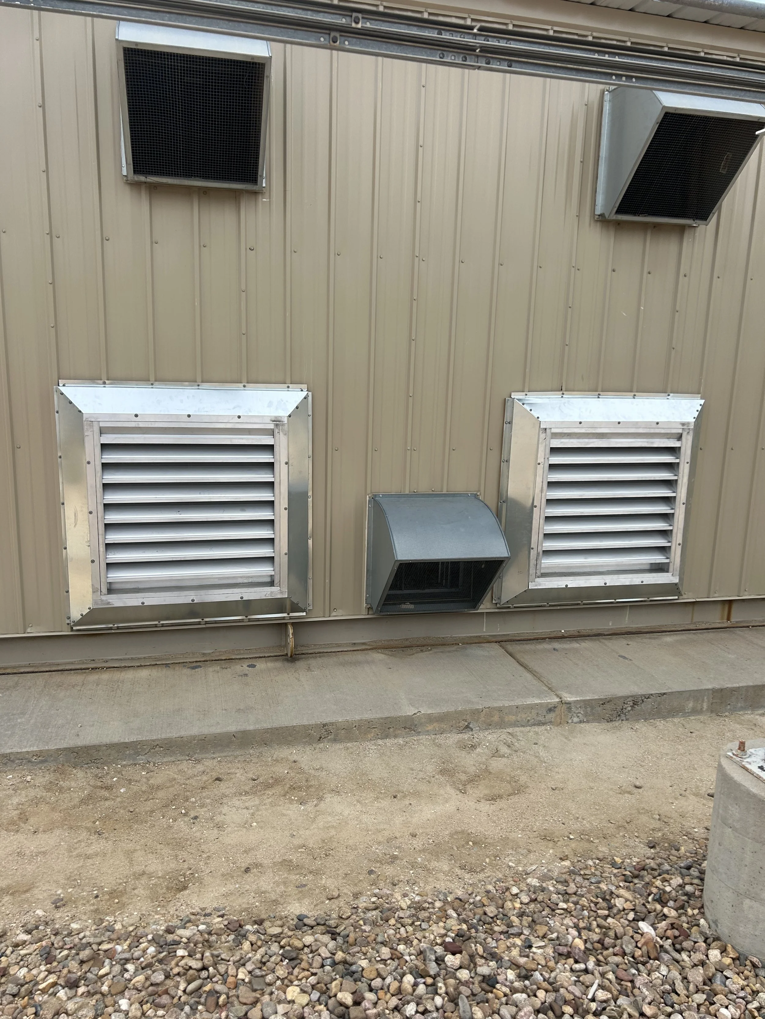 Metal building exterior with four vents and two exhaust fans, with dirt and gravel ground below.