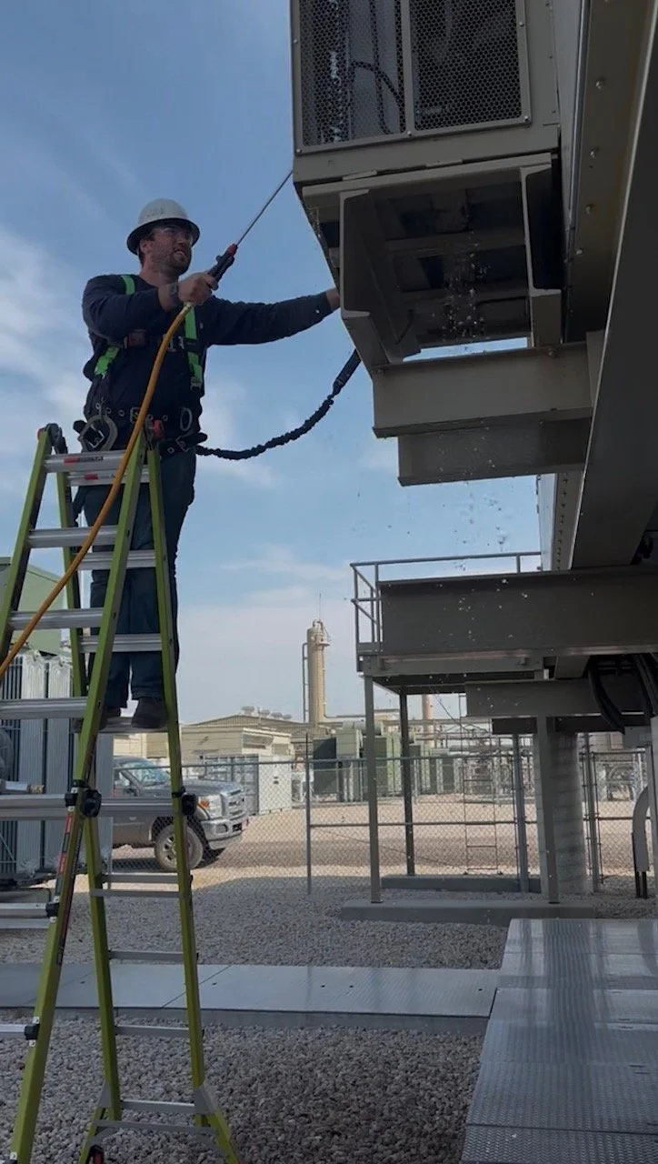 A worker wearing a safety helmet and harness cleaning an industrial air conditioning or ventilation unit using a high-pressure water hose. The worker is safely standing on a ladder outdoors with a clear sky in the background.