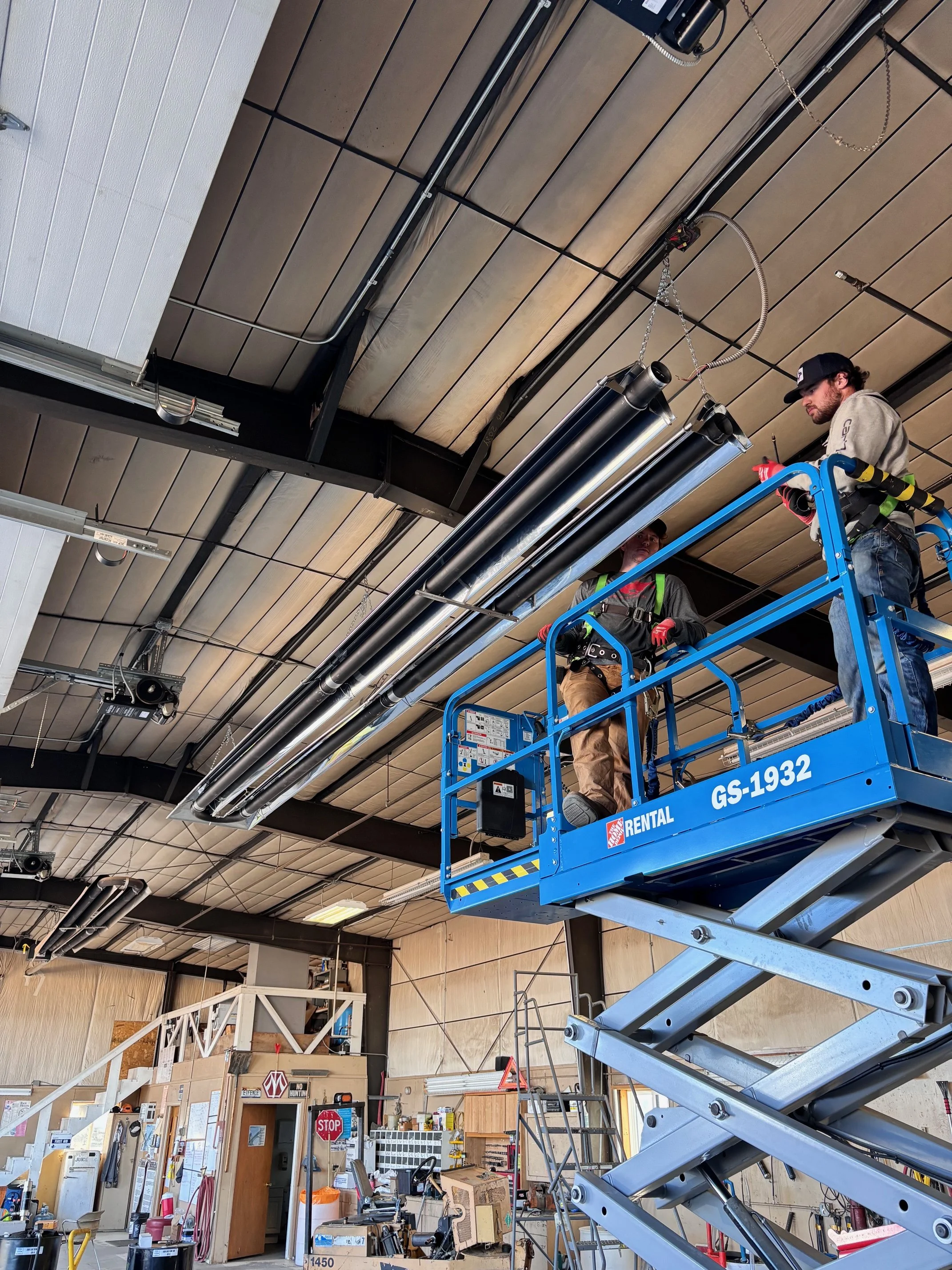 Two workers standing on a blue scissor lift working on a tube heater in a warehouse or industrial space. The ceiling has black metal framework, pipes, and electrical wiring.