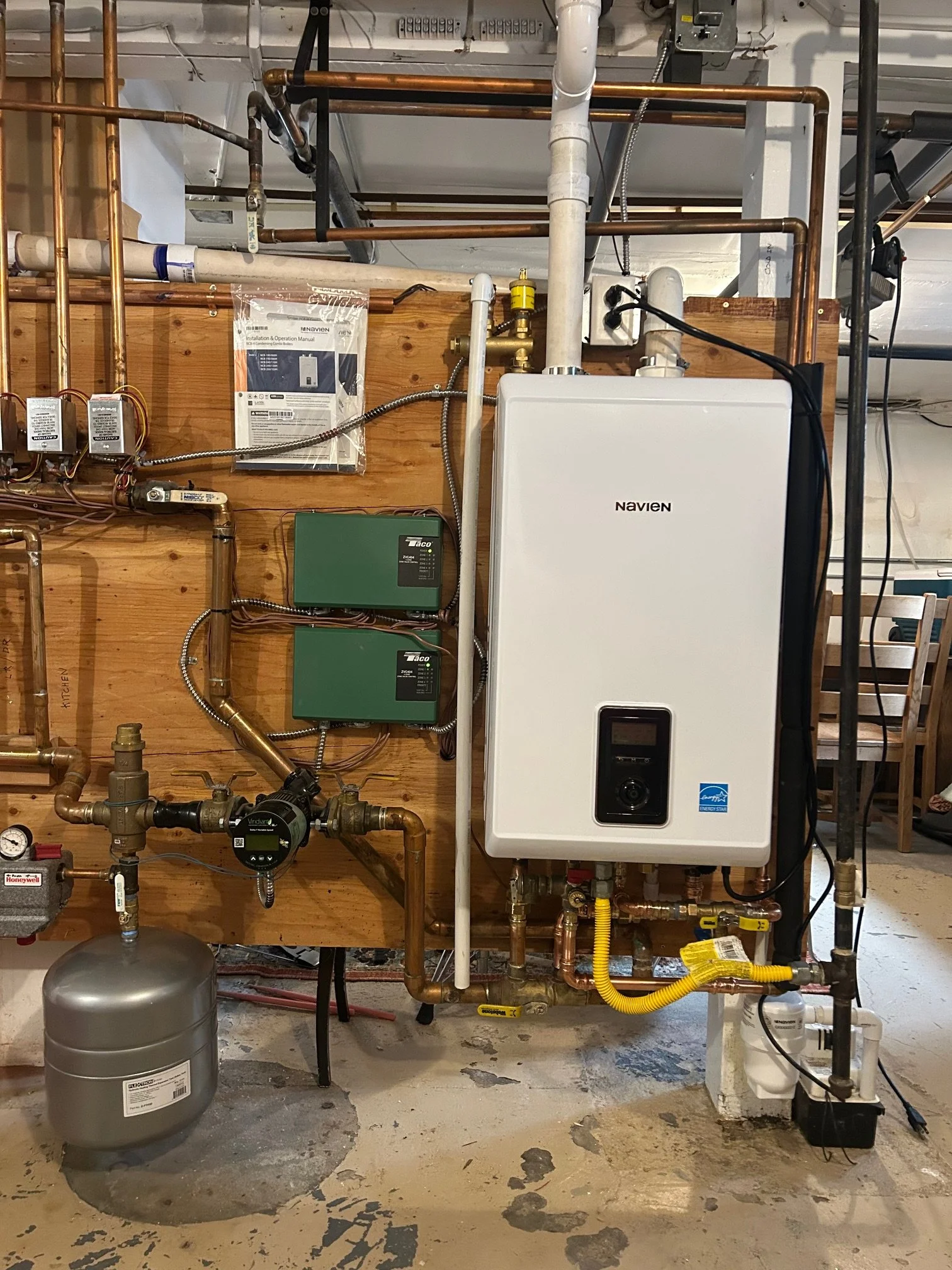 A plumbing and heating system installed in a residential home. Includes pipes, gauges, and control units mounted on a wooden board, with a large white Navien tankless water heater and various copper pipes, valves, and electrical components.