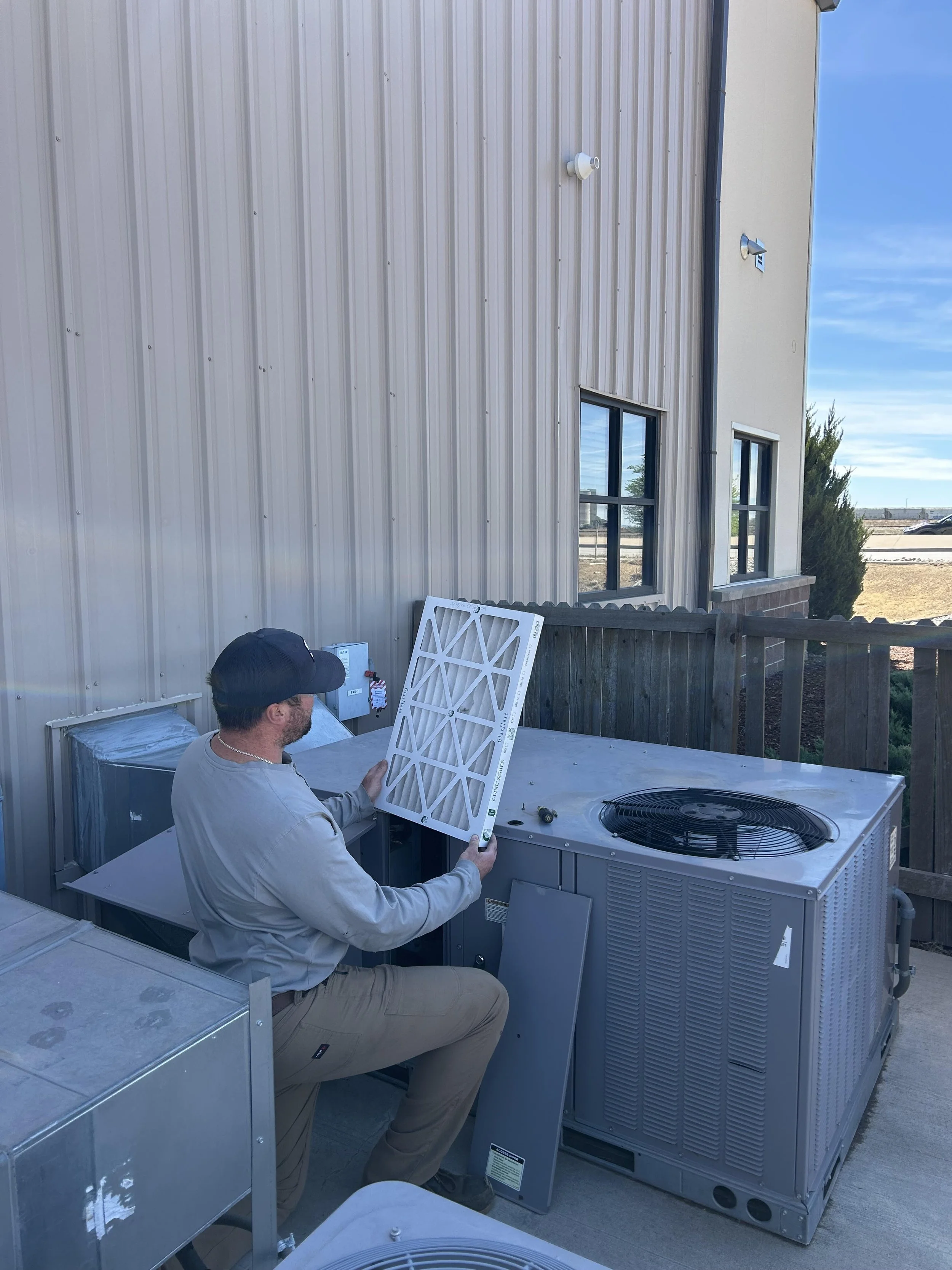 An HVAC system having filters replaced, outside a building with metal siding. He is holding a white pleated filter and there is outdoor equipment and another unit nearby.