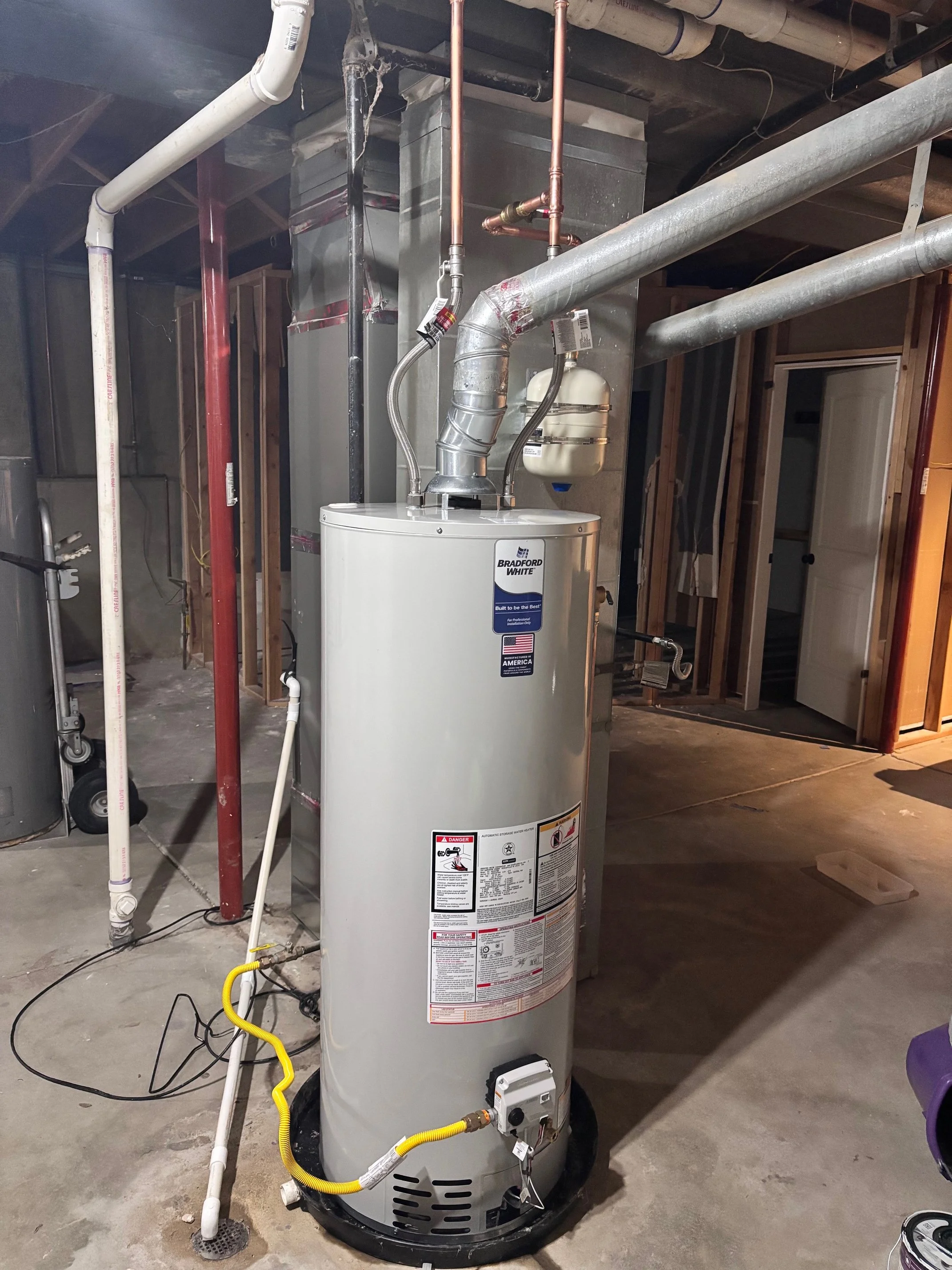 A water heater in an unfinished basement, with pipes and wiring surrounding it.