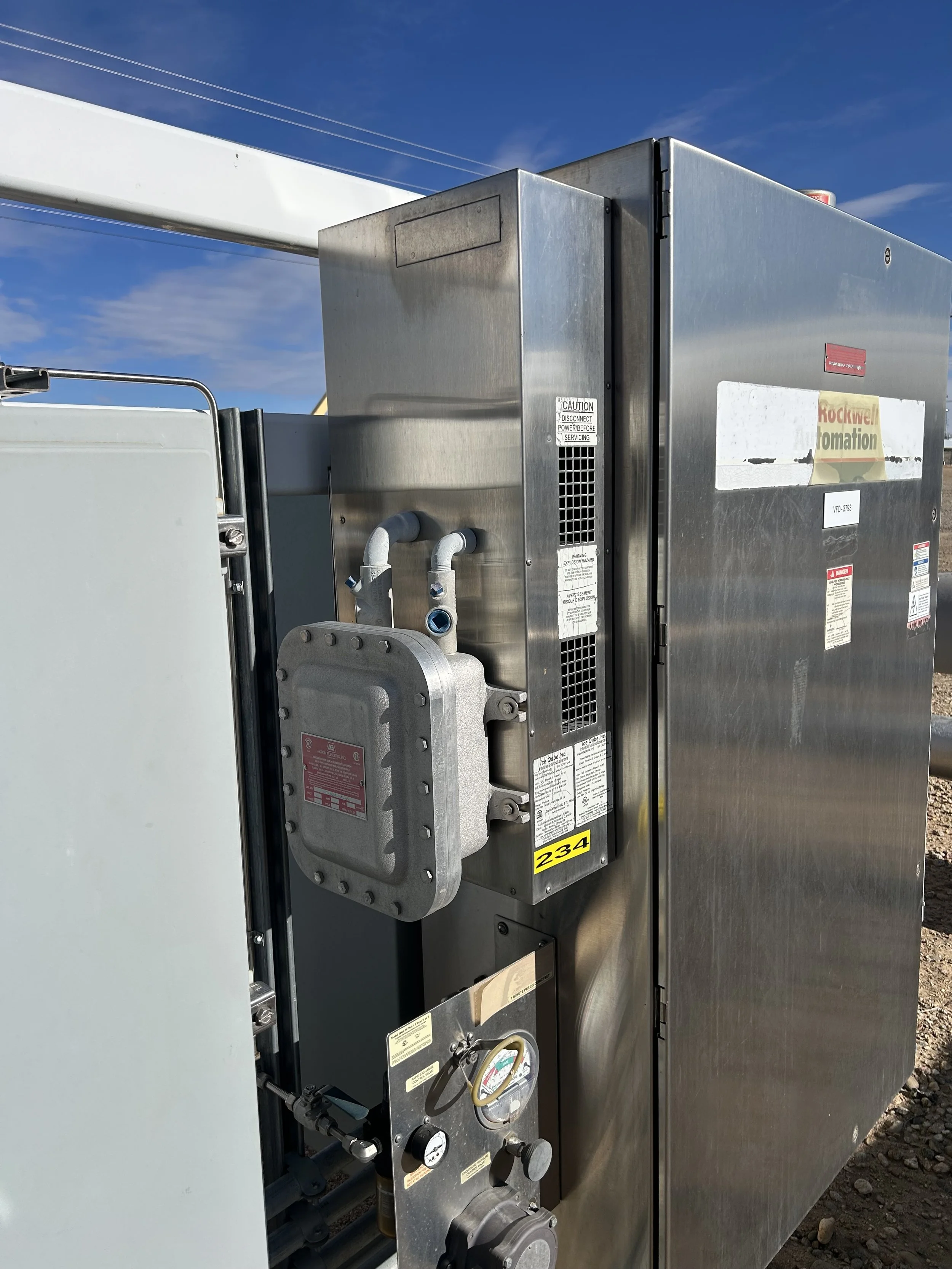 Close-up of industrial electrical equipment with small hazardous location HVAC unit, metal casing, pressure gauges, pipes, and warning labels, outdoors under a blue sky.