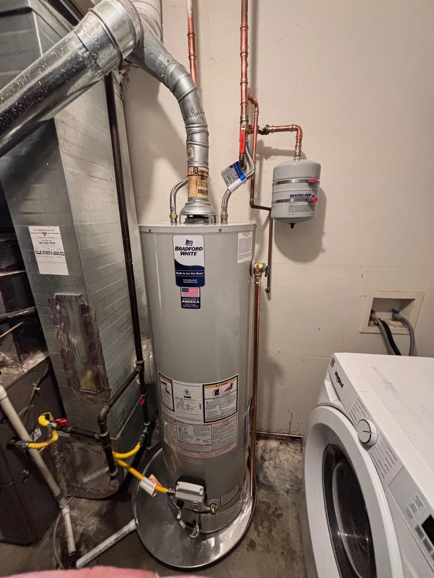 A water heater tank situated in a utility room next to a washing machine. The room appears unfinished with exposed concrete floor and walls. There are plumbing pipes connected to the water heater, and various tools and equipment around.