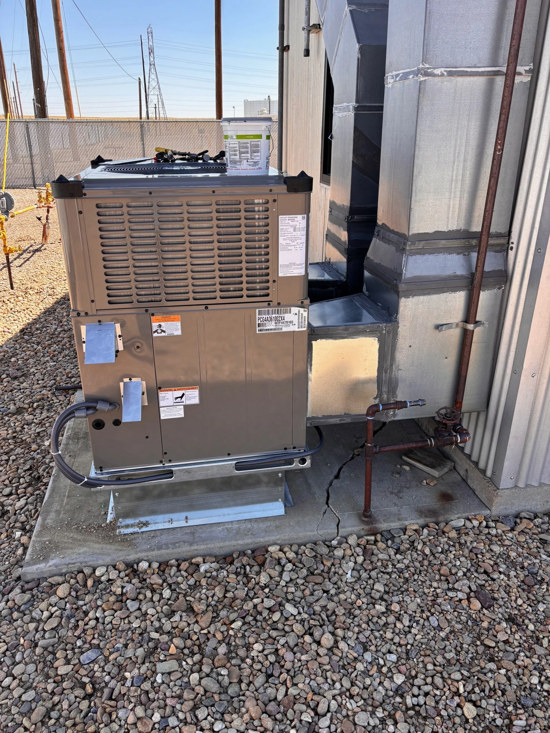 An outdoor air conditioning unit installed on a concrete pad, with a black and silver duct system connected to a building wall. Gravel covers the ground around the unit, and utility poles and power lines are visible in the background under a clear sk