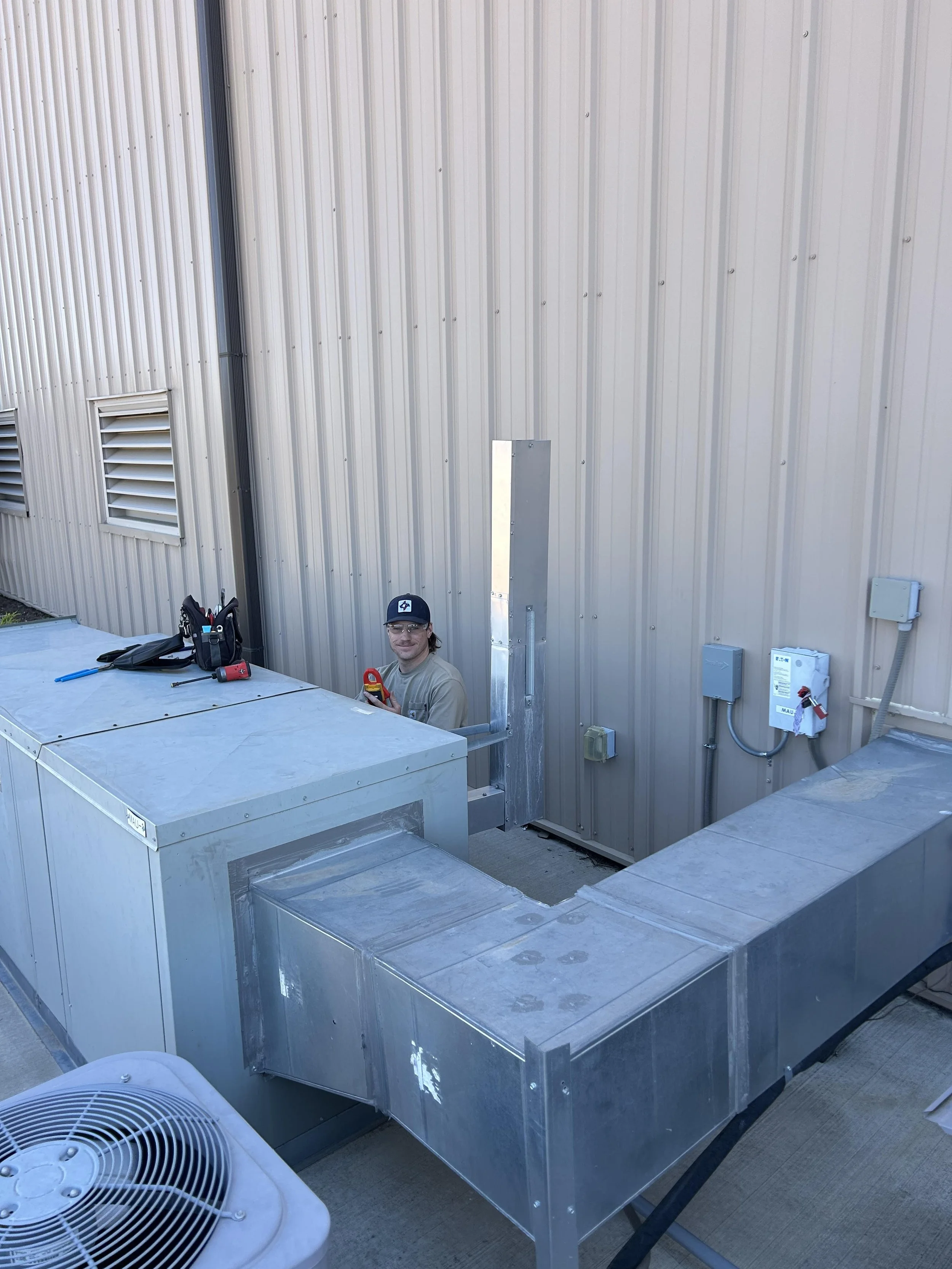 A young man with safety glasses is working outside on a rooftop or industrial area, sitting beside large metallic HVAC units, with tools and a bag nearby, and a LOTO. against a beige metal building wall with vents and electrical boxes.