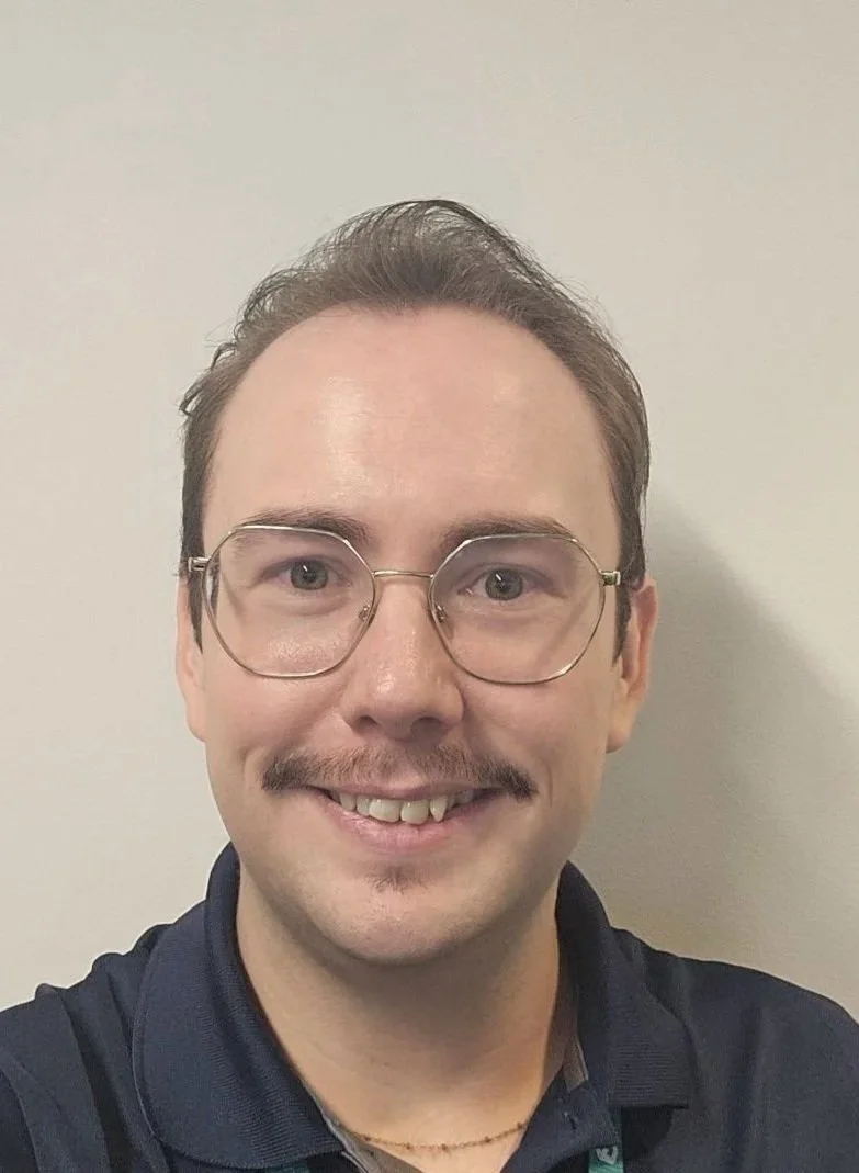 Un hombre con gafas y bigote que sonríe delante de una pared lisa de color claro.