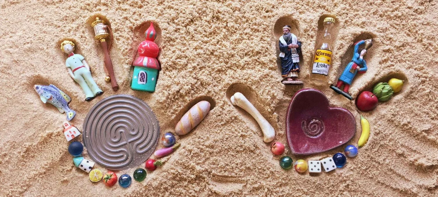 Various small figurines, jewelry, and toys arranged on sand to resemble a heart shape.