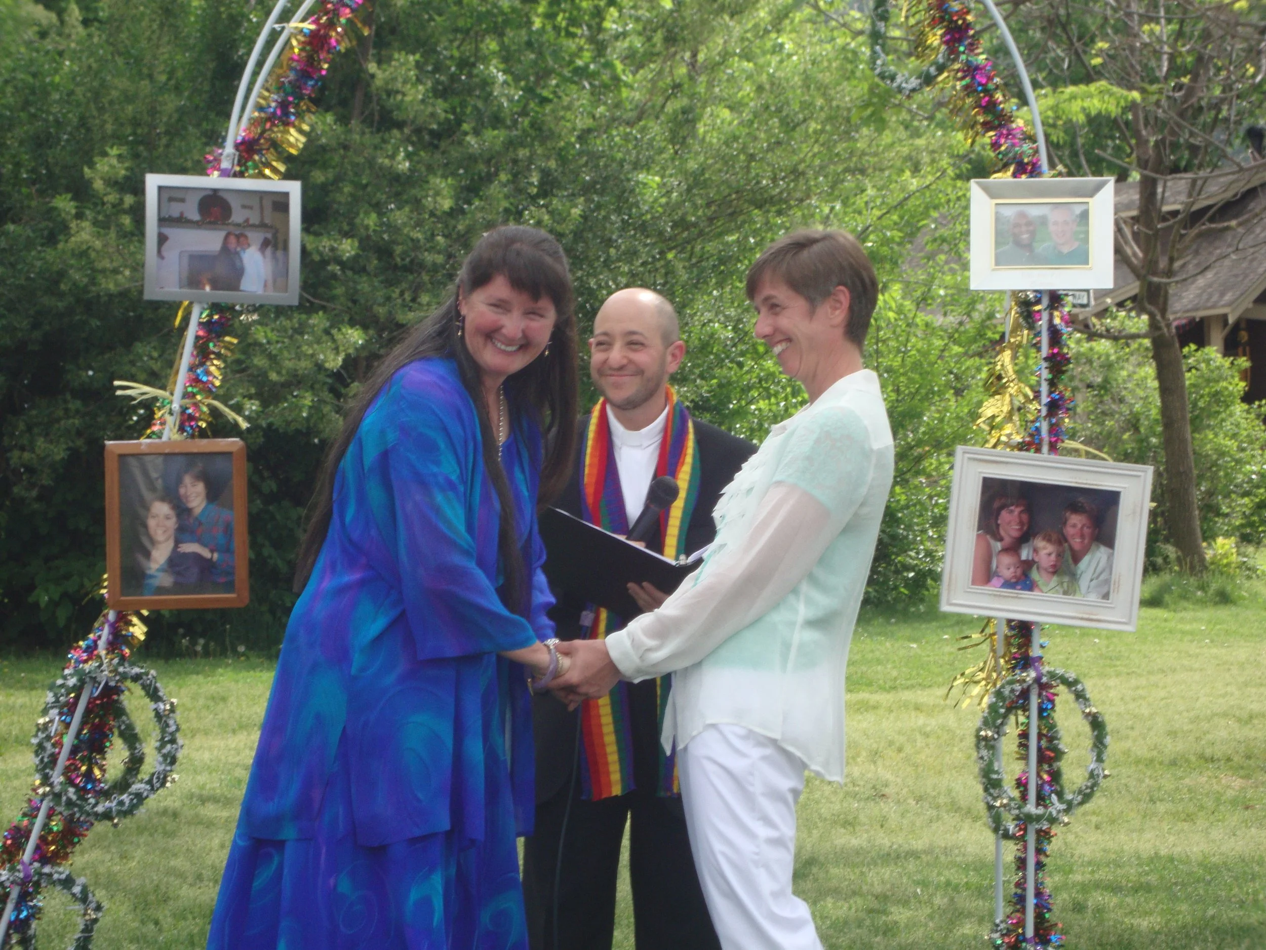 Three people standing in front of a decorated archway during an outdoor celebration, smiling and holding hands, with framed photographs hanging on the arch.