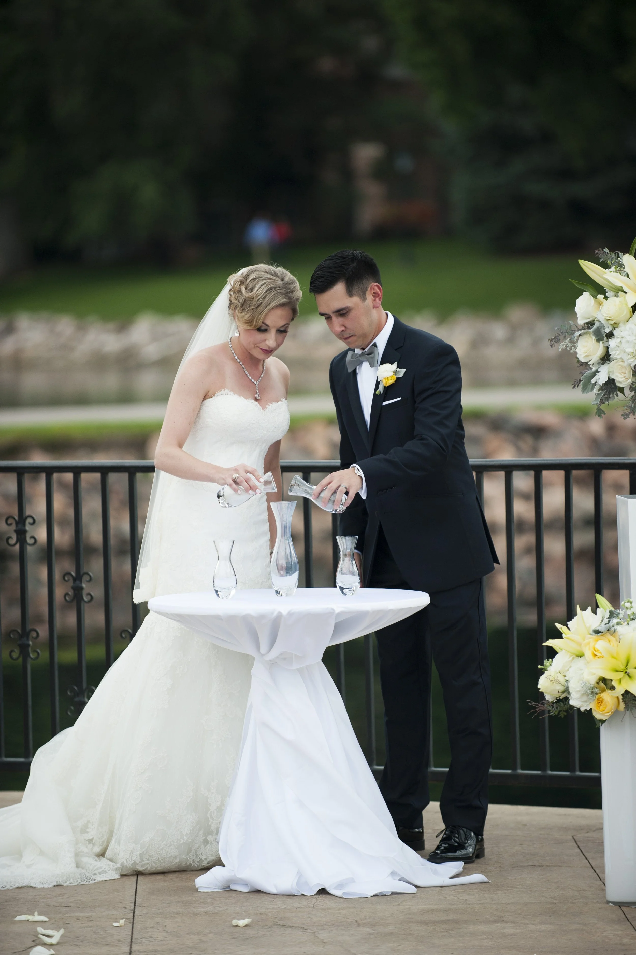 A bride and groom pouring water into cups during their wedding ceremony outdoors.