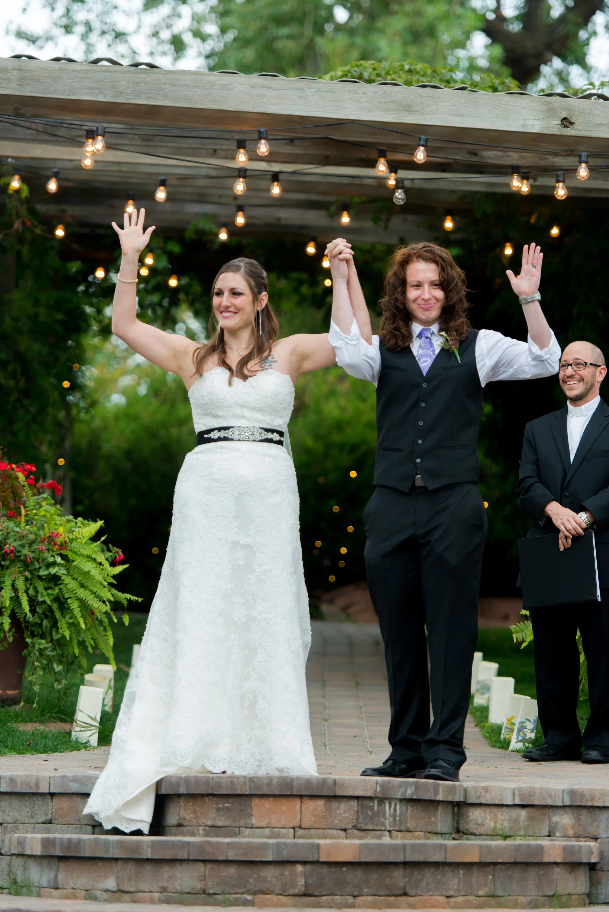 A newlywed couple celebrating their wedding outdoors, holding hands and raising their arms in joy, with an officiant smiling nearby, under string lights and surrounded by greenery.