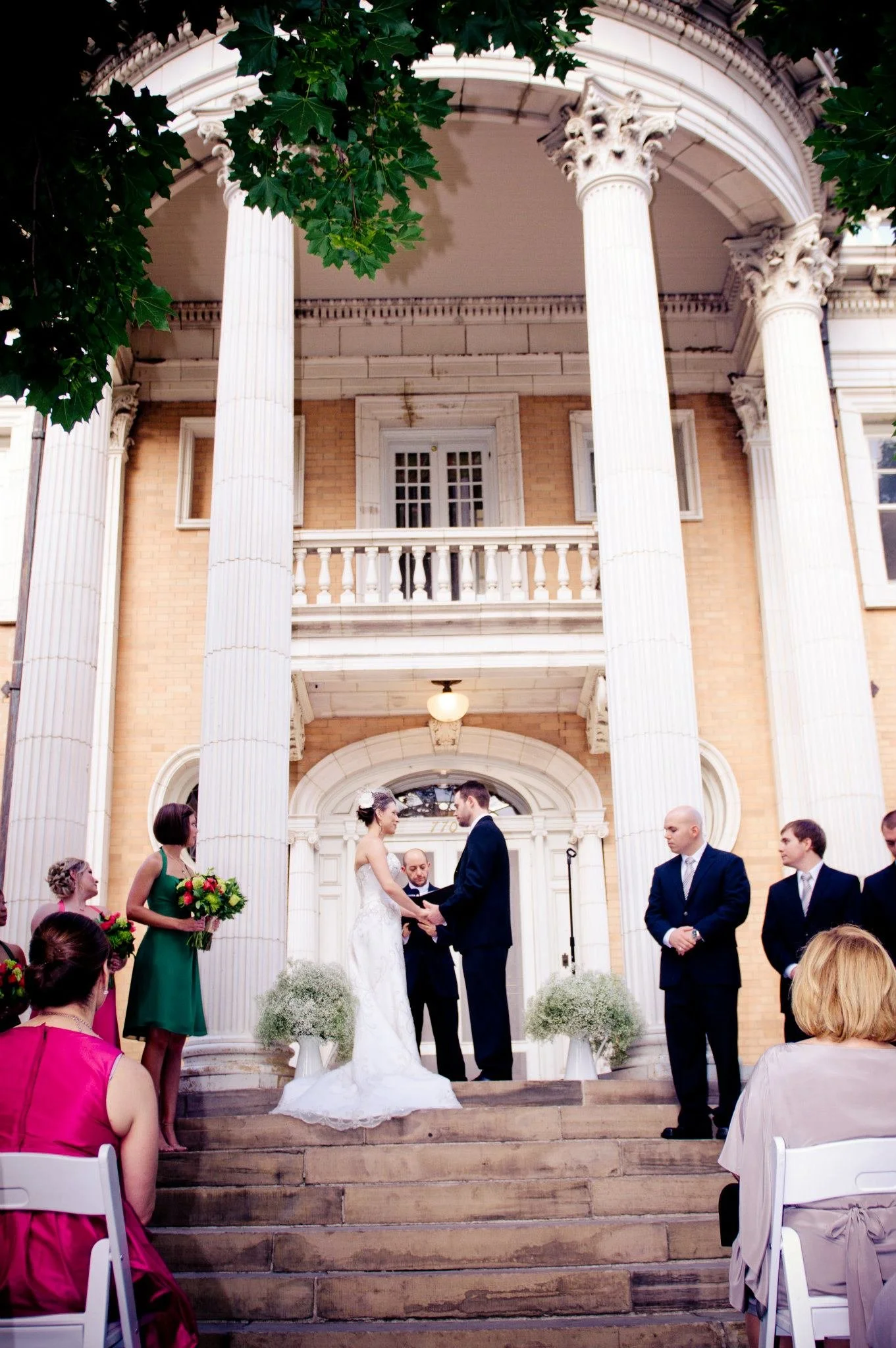 A wedding ceremony taking place on the steps of a historic building with tall white columns and a brick facade. The bride and groom are standing in front of an officiant, surrounded by the wedding party and guests. The bride is wearing a white wedding gown, and the groom is in a dark suit. Bridesmaids and groomsmen are dressed in formal attire, and guests are seated on white chairs, some in colorful dresses.