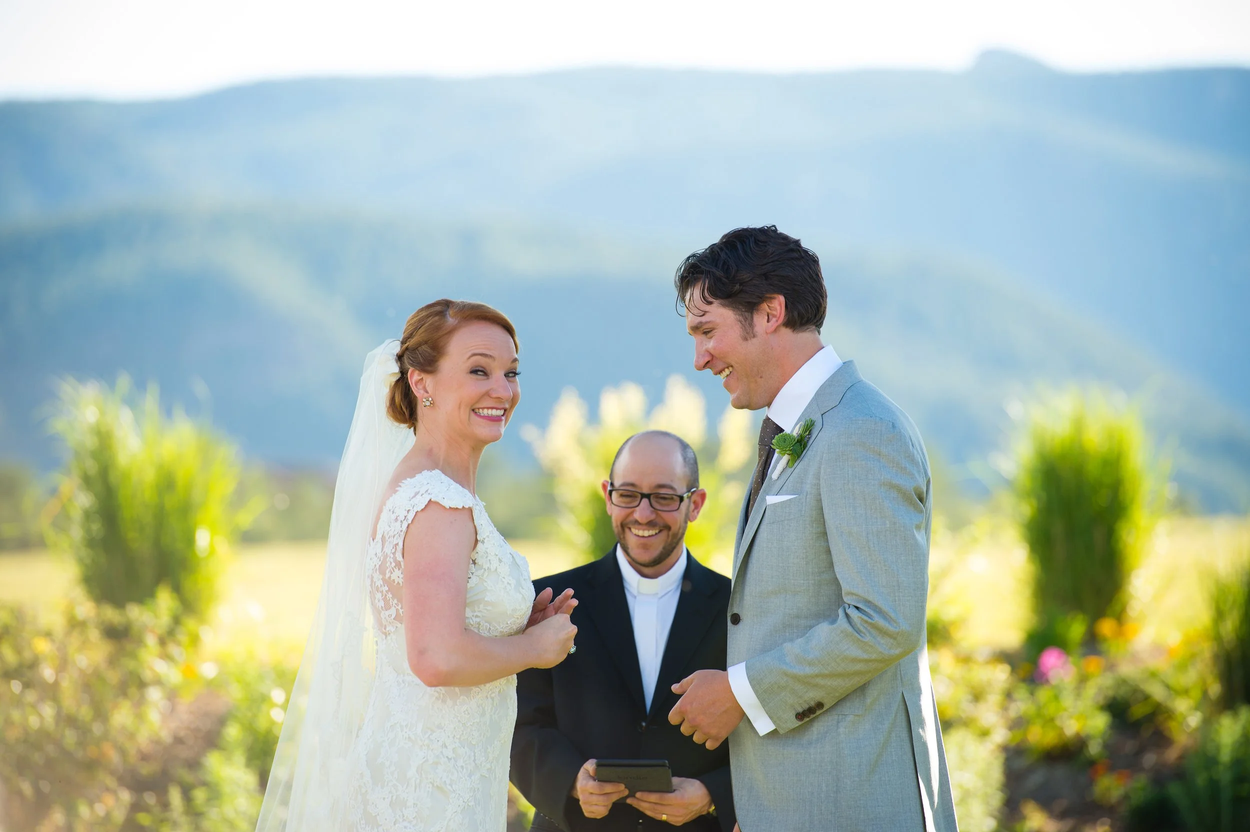 A wedding ceremony outdoors with a bride and groom exchanging vows. The bride is wearing a white lace wedding dress, and the groom is in a light gray suit. There is an officiant holding a tablet, and a scenic mountain landscape in the background.