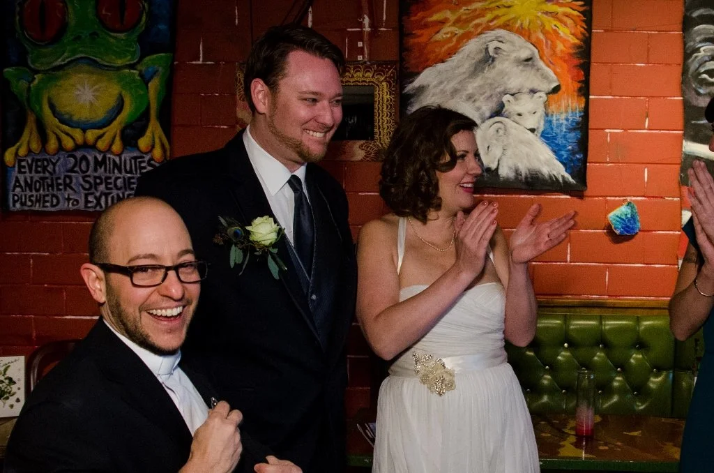 A group of four people at a celebration, with two men and a woman in the center, one of whom is wearing a suit with a boutonniere, and the woman is in a white dress with a decorative belt, clapping and smiling. The setting includes colorful artwork and a brick wall.
