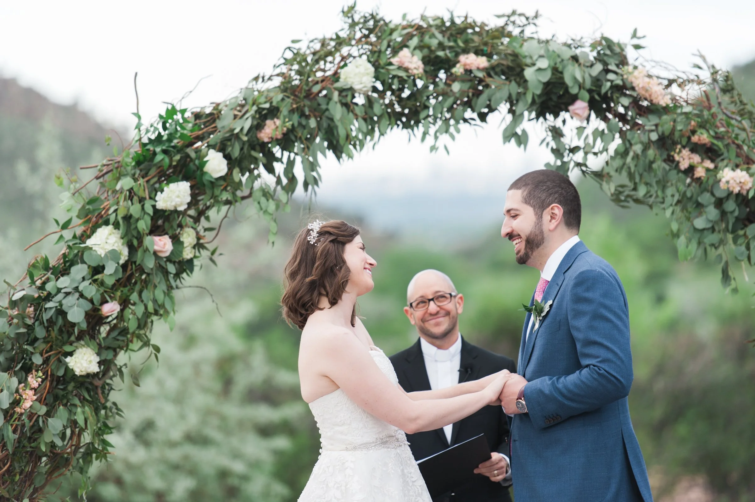 A bride and groom holding hands and exchanging vows during their outdoor wedding ceremony beneath a floral arch with green leaves and pink and white flowers, with an officiant standing behind them.