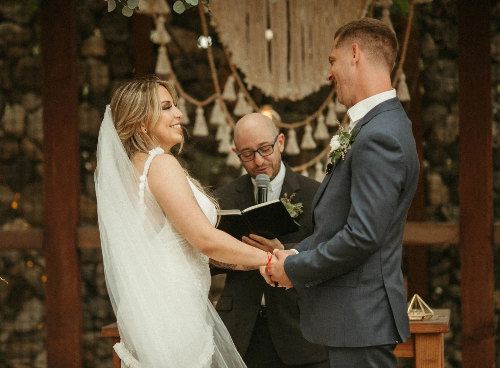 A couple getting married, holding hands, facing each other, smiling, with an officiant reading from a book behind them, in a rustic venue with wooden decor and string lights.