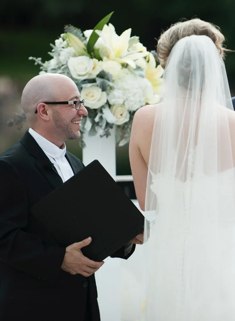 A wedding ceremony with a groom in glasses and a tuxedo smiling at a bride in a white wedding dress and veil. There is a large bouquet of white flowers in the background.