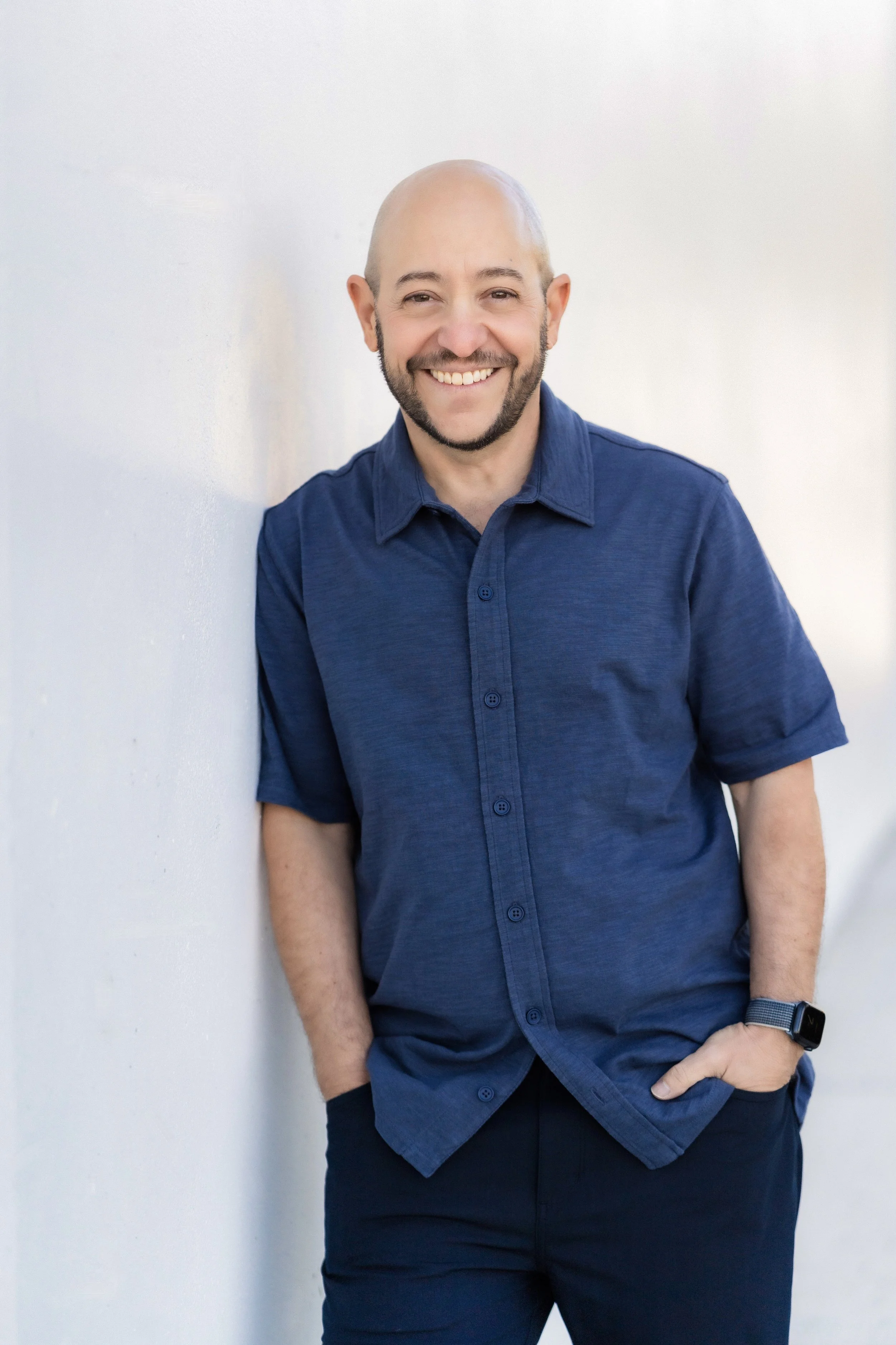 A smiling bald man with a beard wearing a navy blue button-up shirt, standing against a plain white wall with his hands in his pockets.