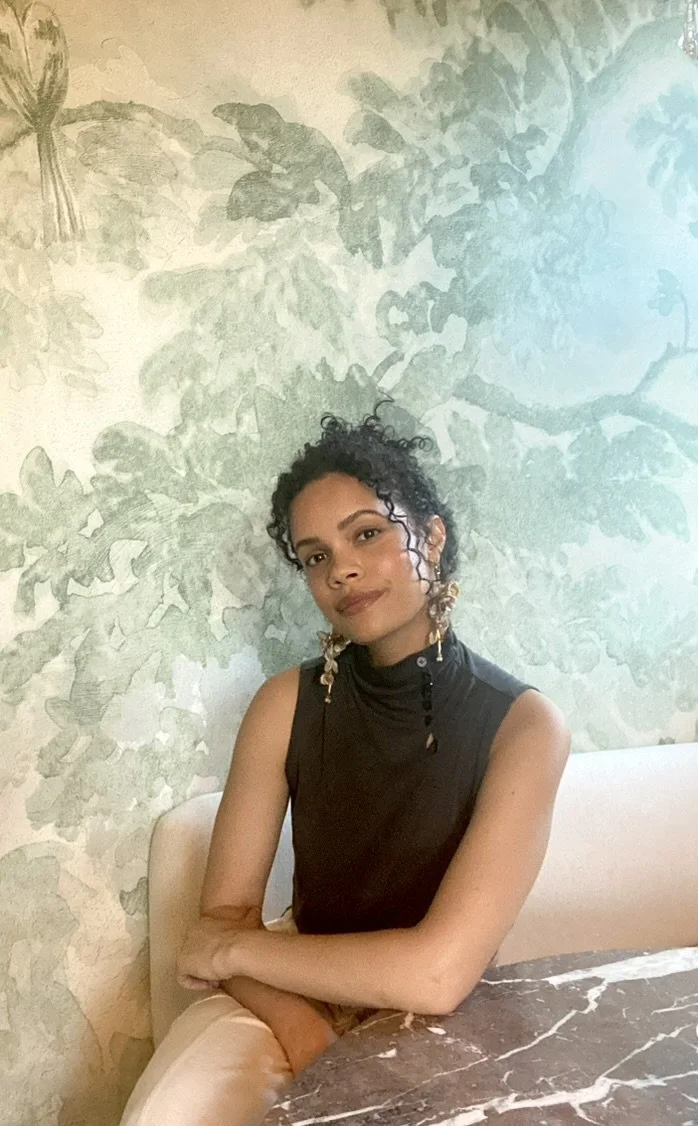 A woman with short curly hair, wearing a black sleeveless top and large earrings, sitting at a table with a marble top against a leafy green mural wall.