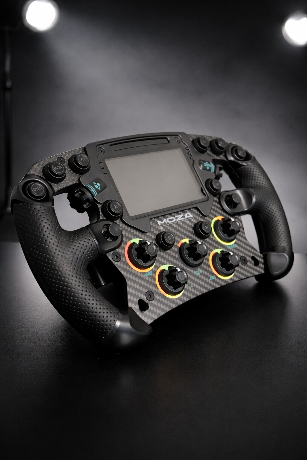 A racing car steering wheel with multiple buttons and knobs, made by MOZA, placed on a dark surface with stage lights in the background.