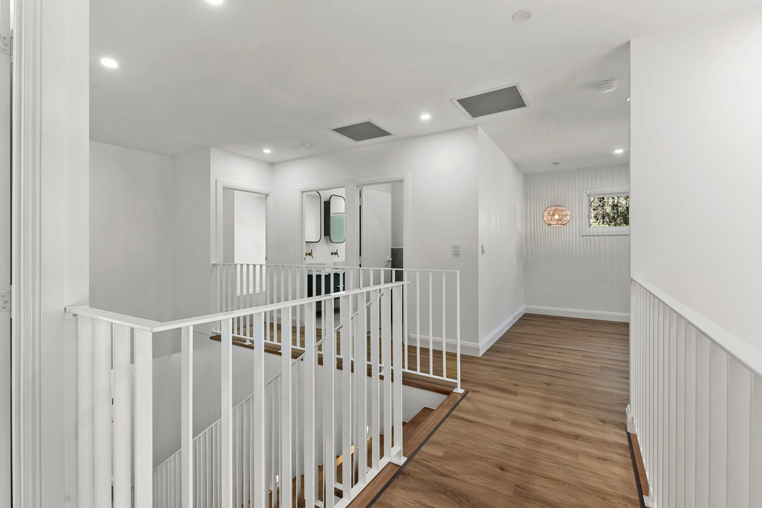 A bright, modern indoor space with white walls, wooden flooring, and ceiling lights. There is a staircase with white railings, a small window with a view of trees outside, and a pendant light fixture.