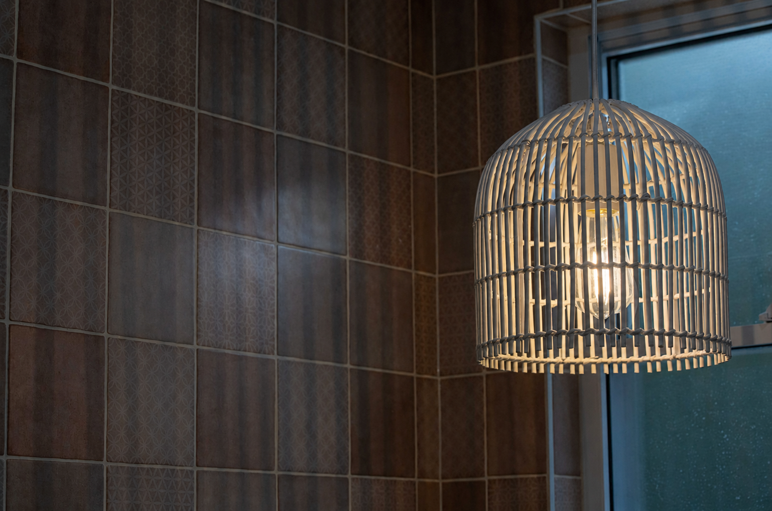 A wicker pendant light hanging in a bathroom near a tiled wall and a window, with the light turned on.