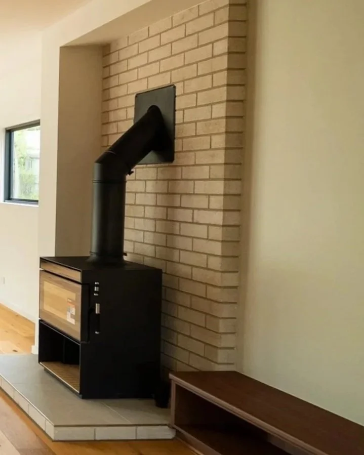 A black wood stove with a chimney pipe extending through a rectangular black vent in a brick wall. The stove is on a tiled platform in a room with hardwood floors and a window visible on the left.