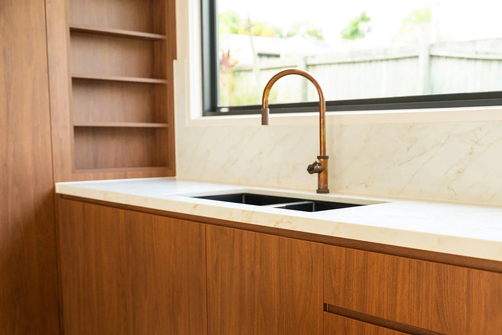 Kitchen with wooden cabinets, a white marble countertop, a copper-colored faucet, and a window overlooking a backyard with a wooden fence and green trees.