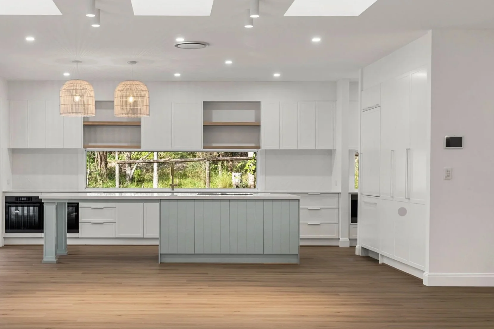 Modern white kitchen with a large window overlooking a wooded outdoor scene, two hanging rattan pendant lights, and wood flooring.