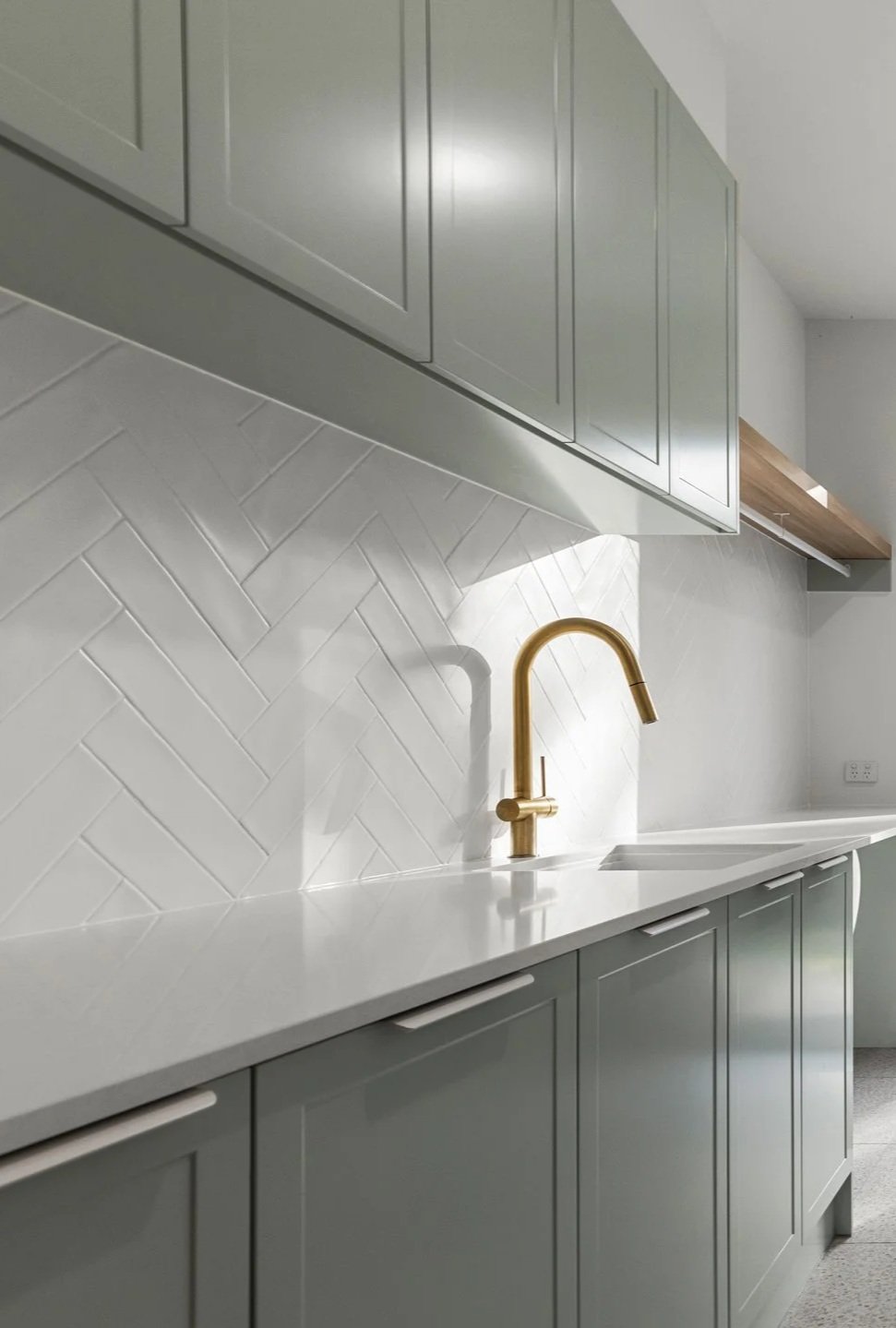 Modern kitchen with light gray cabinets, white countertop, white textured backsplash, and a gold faucet.