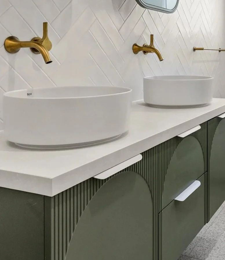 Double white vessel sinks with gold faucets on a white countertop in a modern bathroom with a geometric tile wall and a green cabinet below.