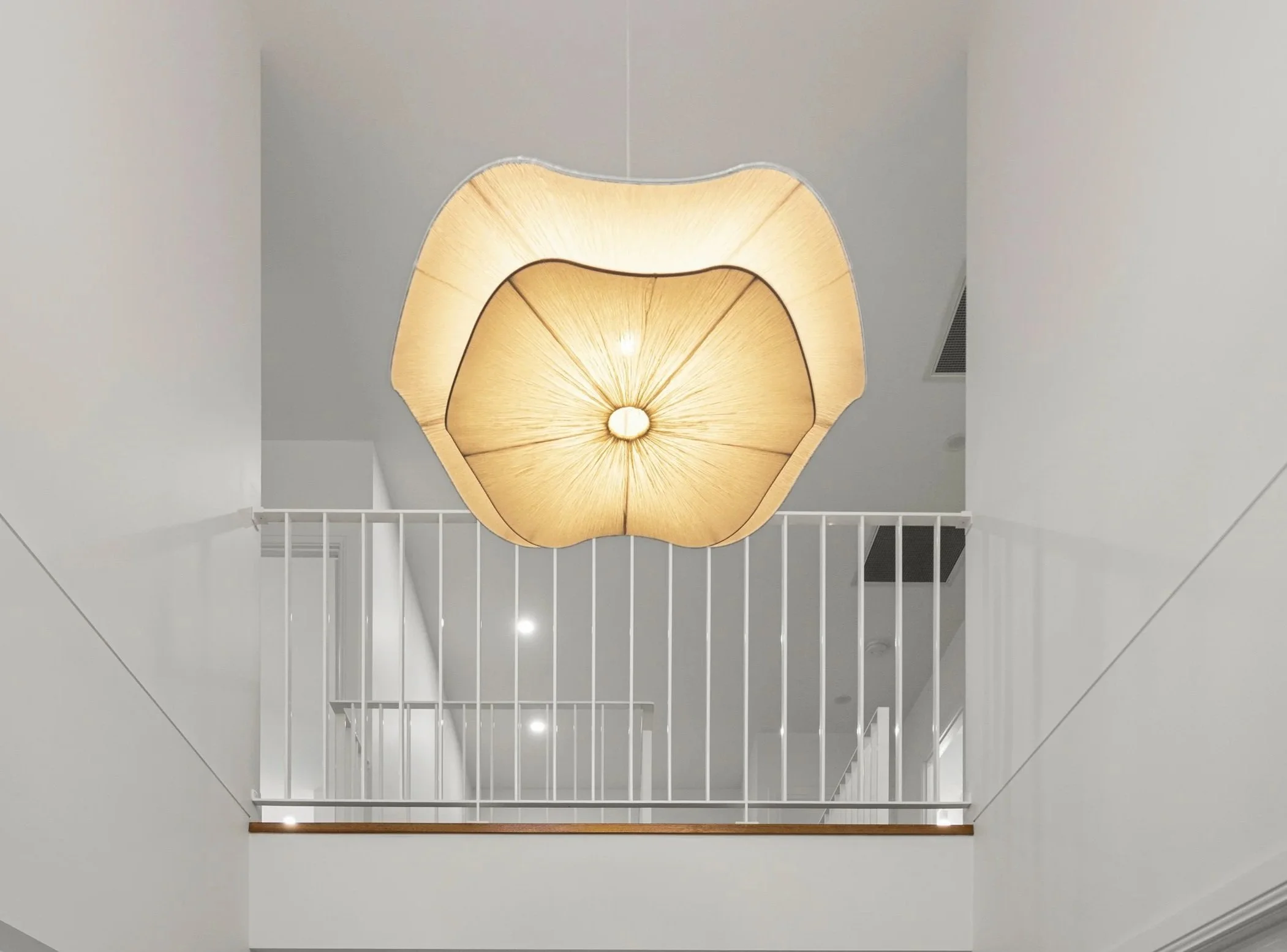 Upward view of a modern chandelier resembling a flower with soft yellow lighting, hanging over a white railing and staircase in a minimalist interior.