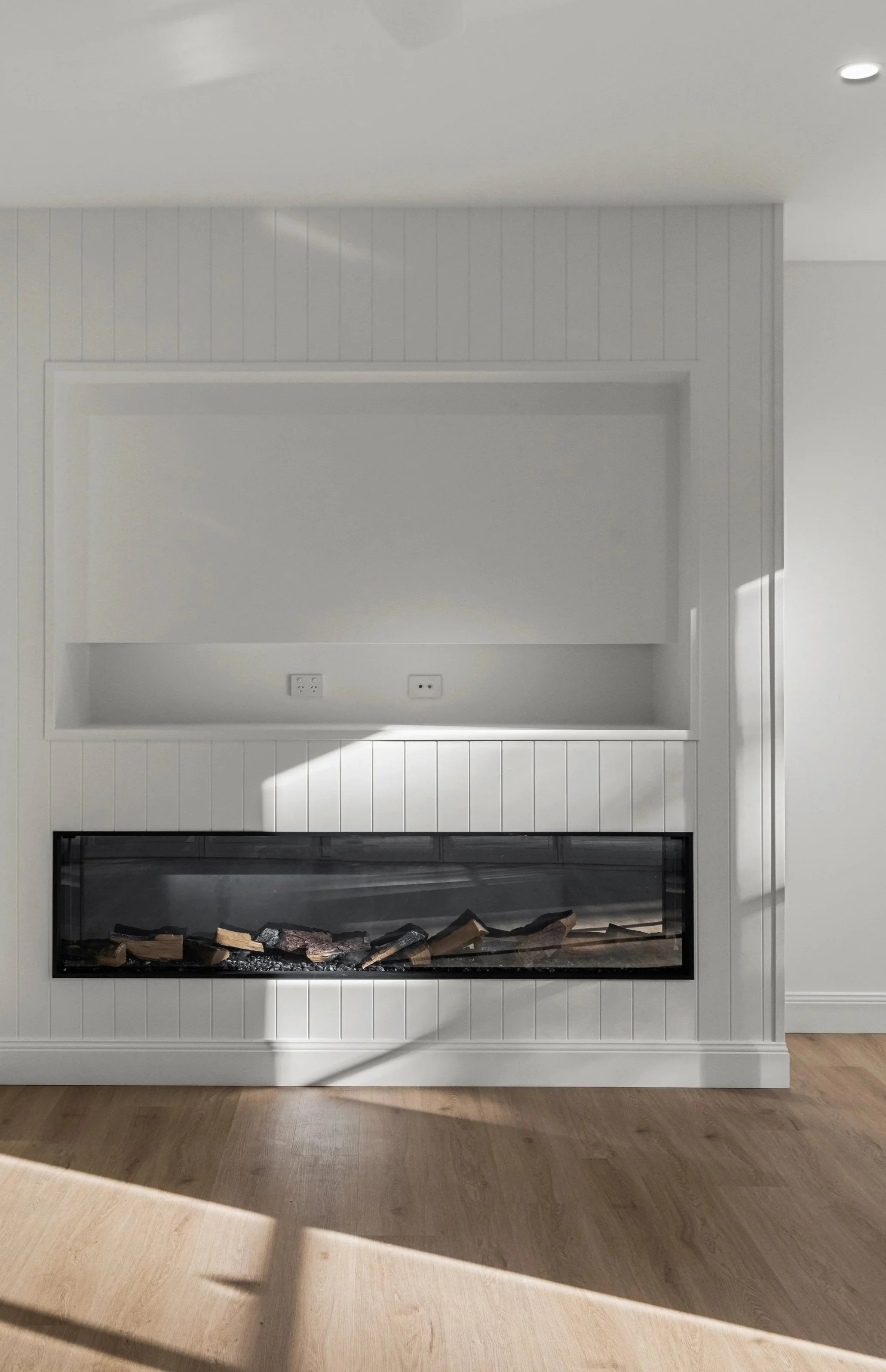 A modern white fireplace with a glass front and logs inside, set in a white-paneled wall in a room with wooden flooring.