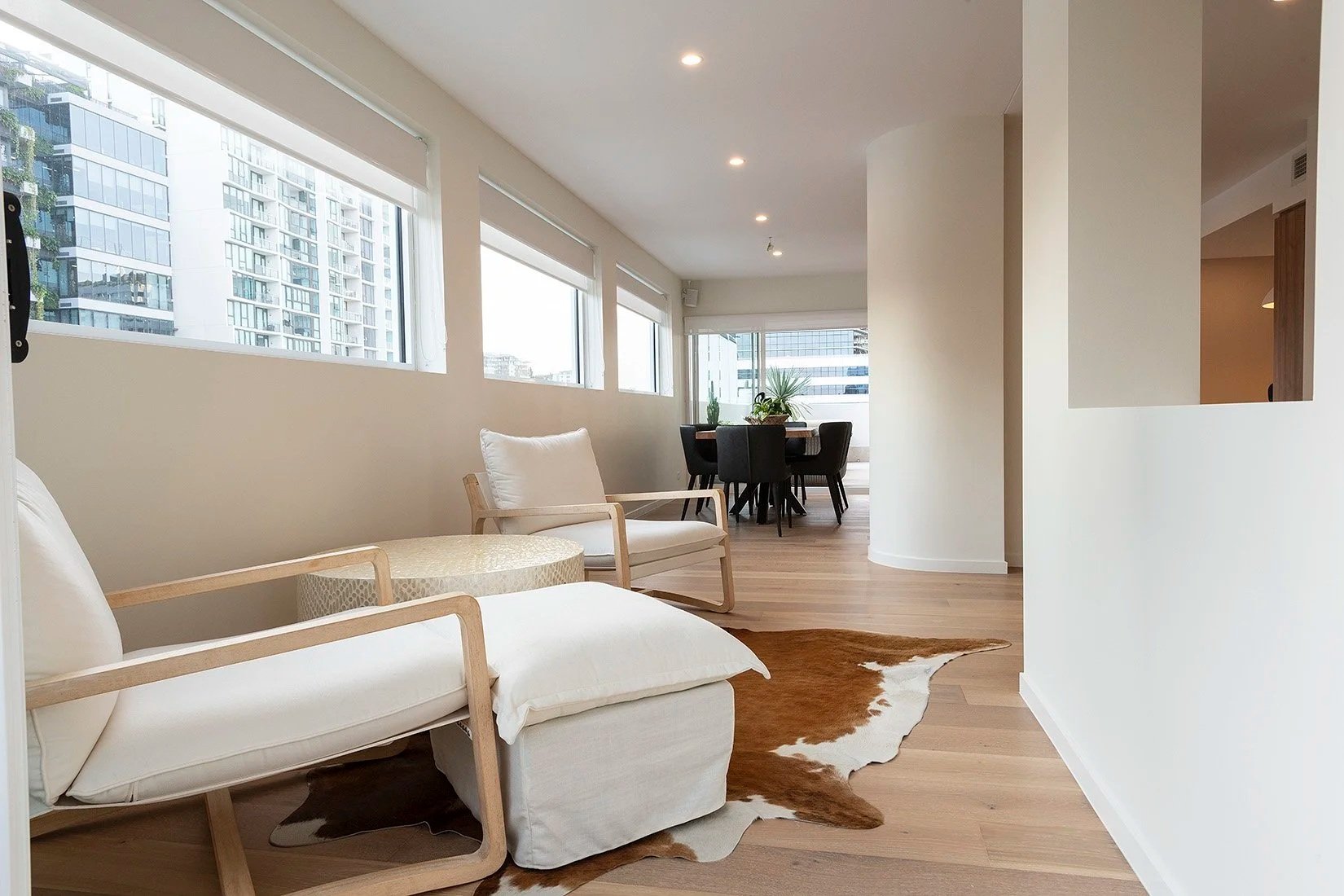 Modern living room with large windows, white armchair with wooden frame, cowhide rug, and dining area with black chairs and a round table with plants.