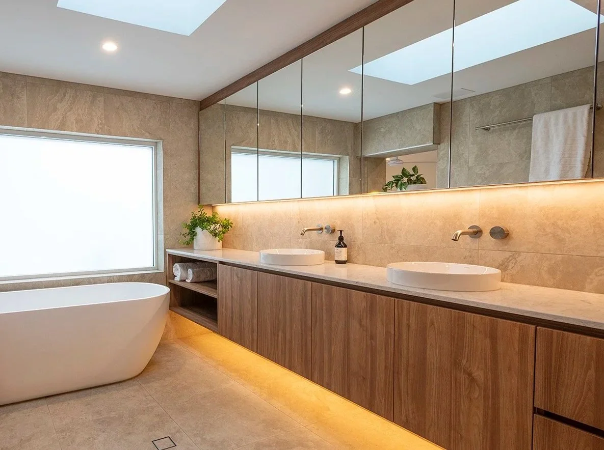 Modern bathroom with a freestanding bathtub, a long wooden vanity with dual vessel sinks, large mirrors, a skylight, and beige tile walls and floor.
