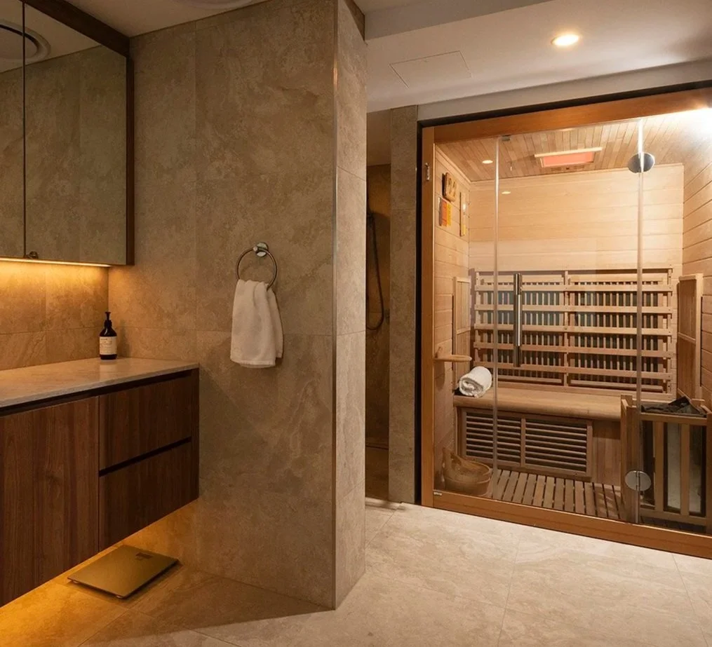 A bathroom with beige tiled walls and floor, a wooden vanity with a beige countertop, a soap dispenser, a round towel ring with a white towel, and a glass-door sauna with wooden interior and a folded white towel inside.